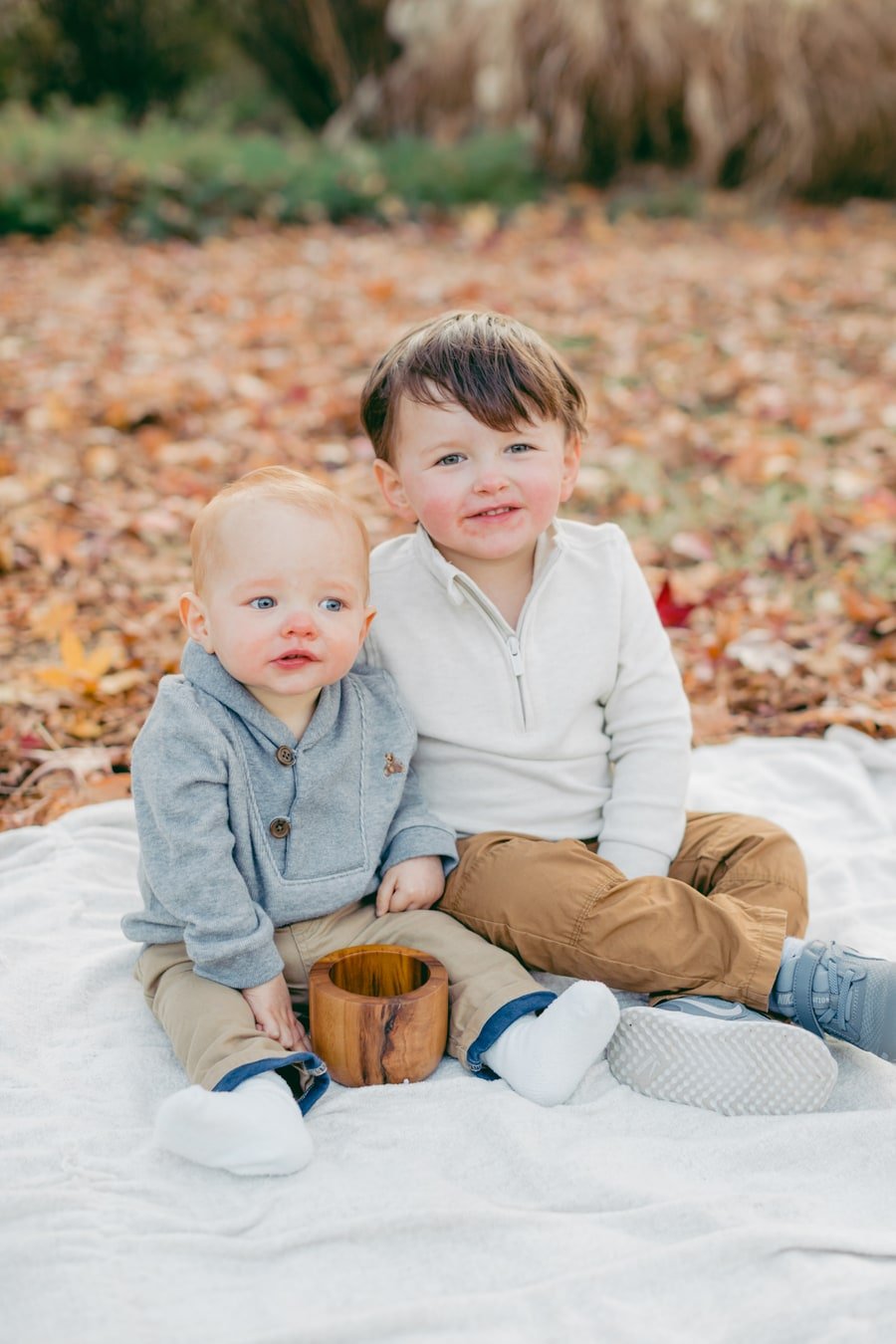 Two young siblings sitting together on a white blanket among fall leaves.