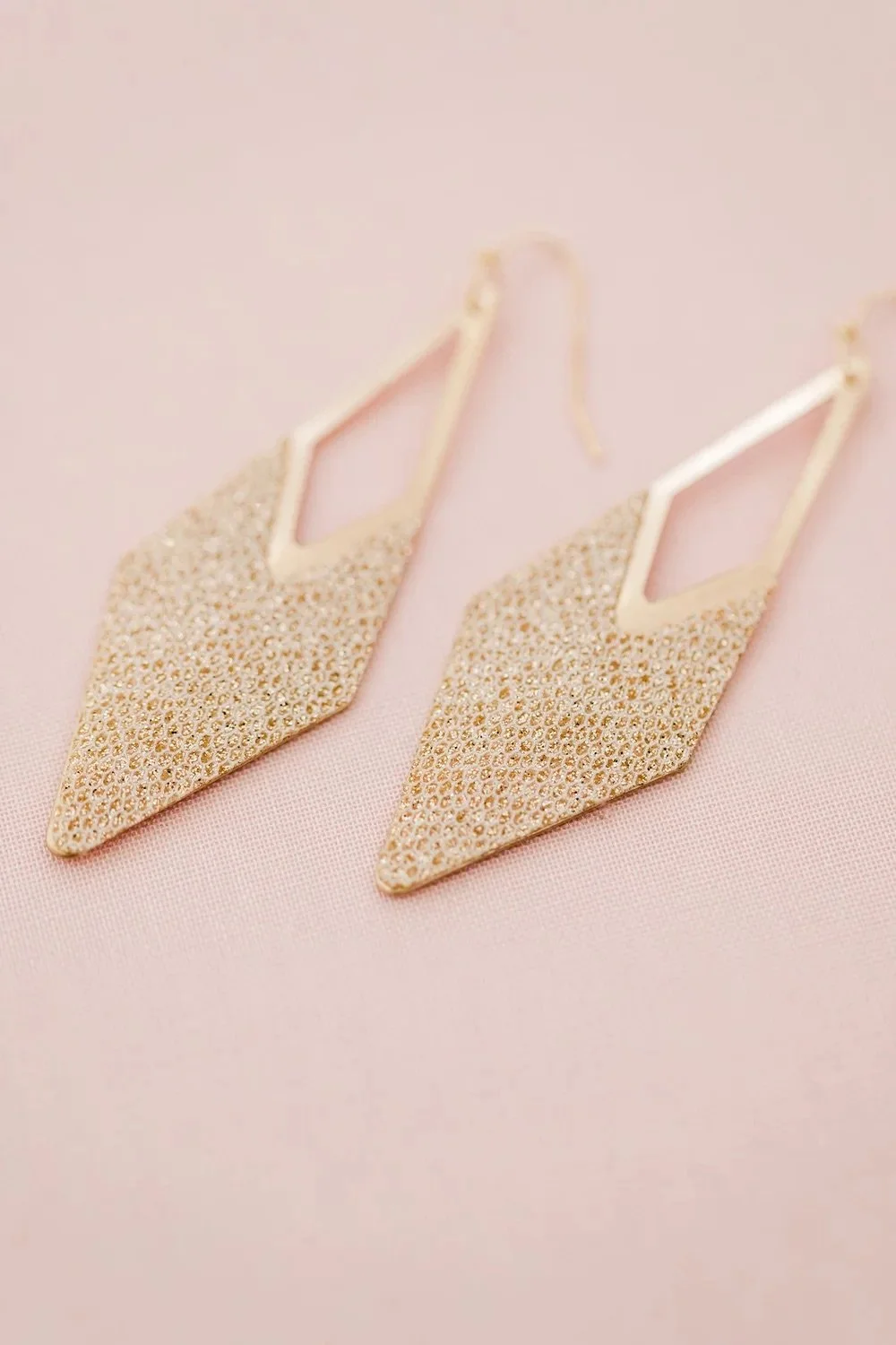 Gold diamond-shaped drop earrings rest on pink fabric.