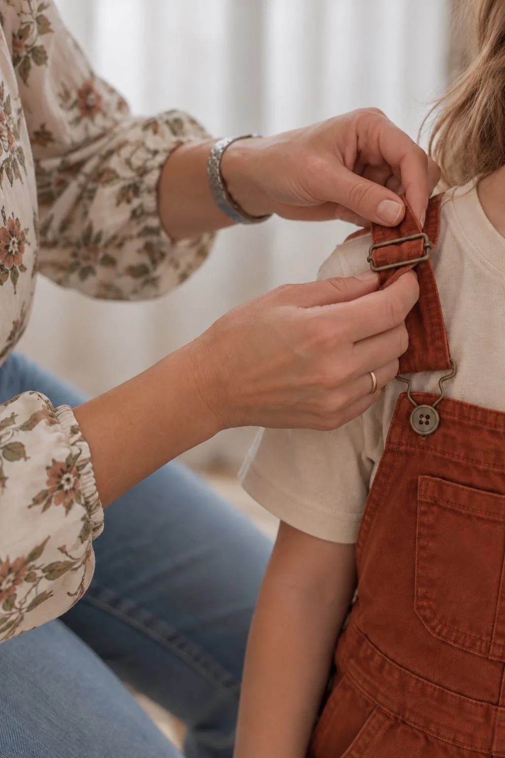 Hands adjusting the strap on a child’s rust overalls over a cream tee.
