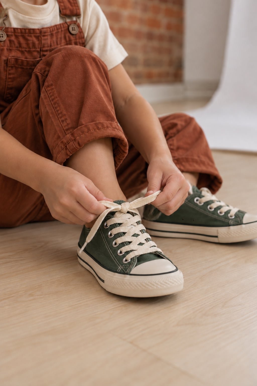 A child in rust overalls tying cream laces on green canvas sneakers.