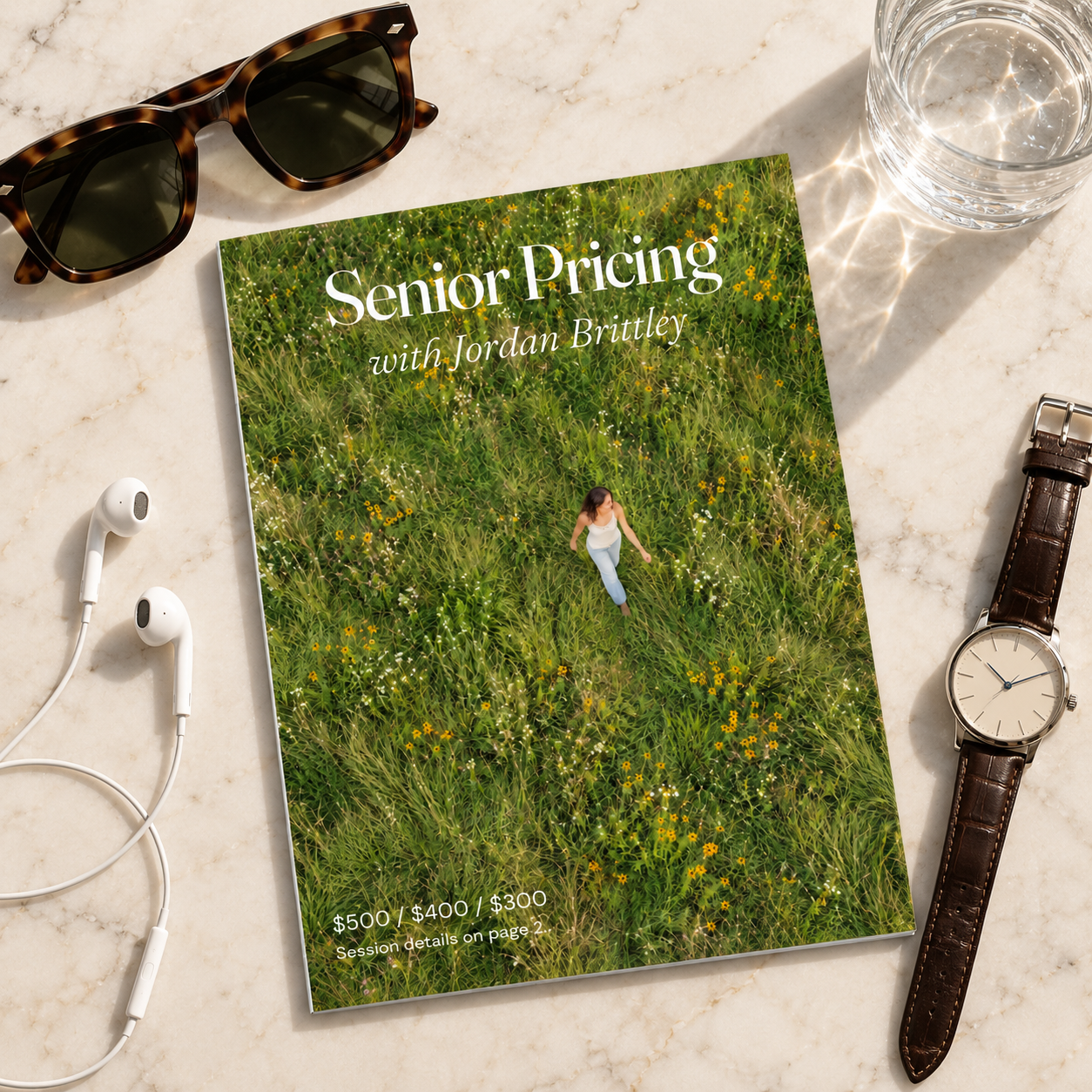 Senior Pricing with Jordan Brittley magazine on marble beside water, tortoiseshell sunglasses, white wired earbuds, and a leather-strap watch.