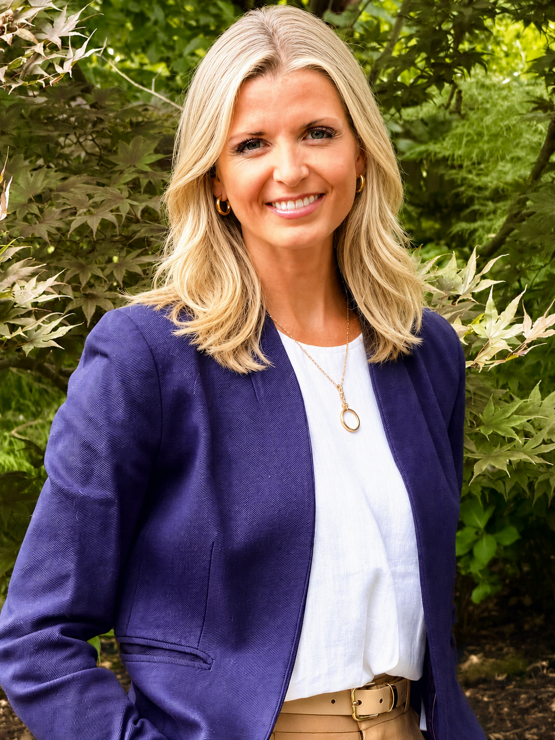 A woman with blonde hair smiling outdoors in front of greenery, wearing a navy blazer, white blouse, gold jewelry, and beige pants.