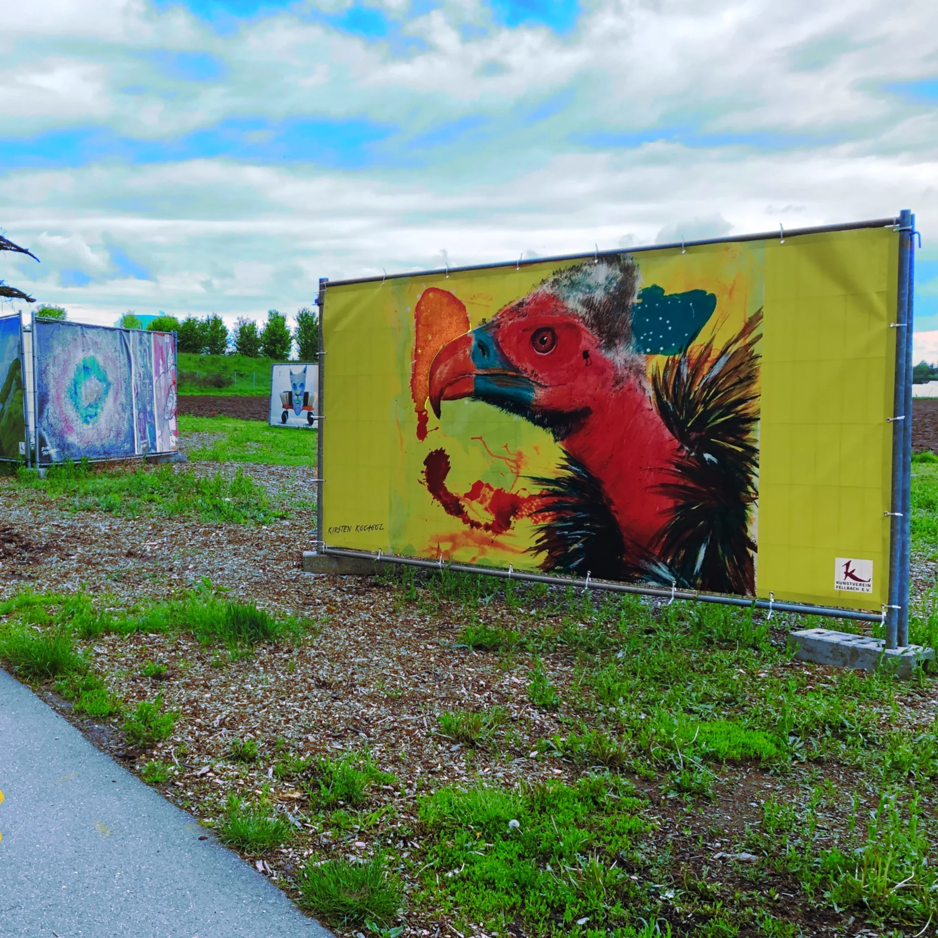 Kunstwerke im Freien, darunter ein großes Bild eines bunten Vogels auf einer gelben Plane. Das Kunstwerk zeigt einen Rotkopf, wahrscheinlich eines Greifvogels, in einem stilisierten und lebhaften Stil.