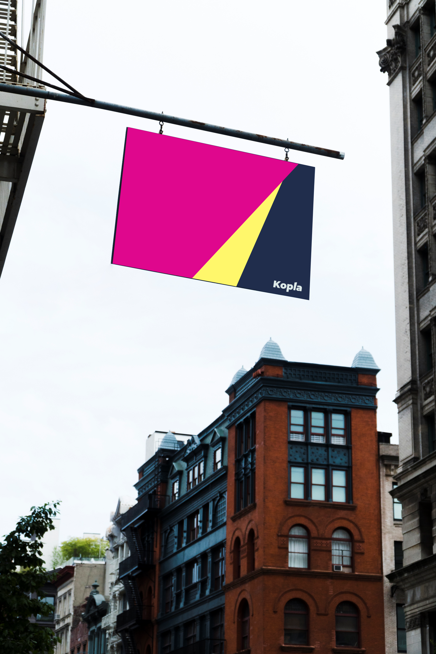 Valokuva katumaisemasta, jonka etualalla roikkuu myymälän kyltti. Kyltissä on Kopla Kollektiivin räväkät värit pinkki, keltainen ja sininen sekä teksti Kopla. Kyltti erottuu realistisesta taustasta räikeydellään.