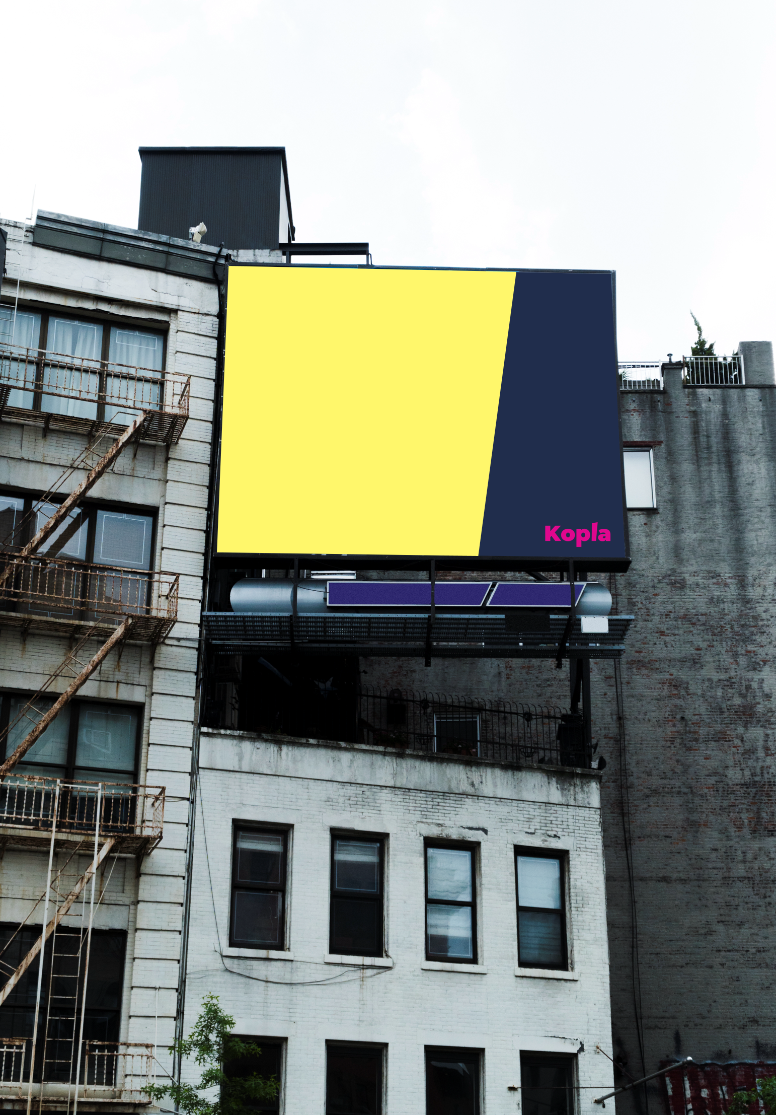 Valokuva ränsistyneestä rakennuksesta, jonka seinustalla on billboard. Billboardilla on Kopla Kollektiivin värit keltainen ja tummansininen ja alareunassa teksti Kopla pinkillä
