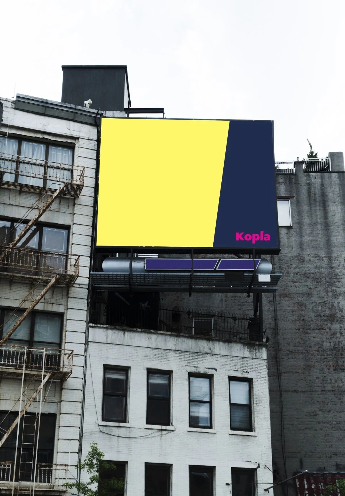 Valokuva ränsistyneestä rakennuksesta, jonka seinustalla on billboard. Billboardilla on Kopla Kollektiivin värit keltainen ja tummansininen ja alareunassa teksti Kopla pinkillä