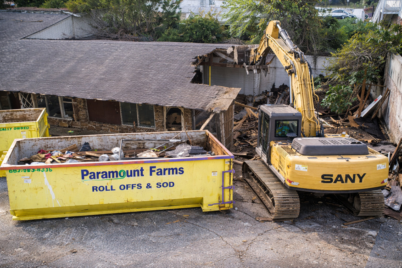 Hernando MS Demolition Job showing our dumpster. A yellow demolition excavator in operation, tearing down a building with debris scattered around, and a bright yellow dumpster labeled 'Paramount Farms' filled with trash.