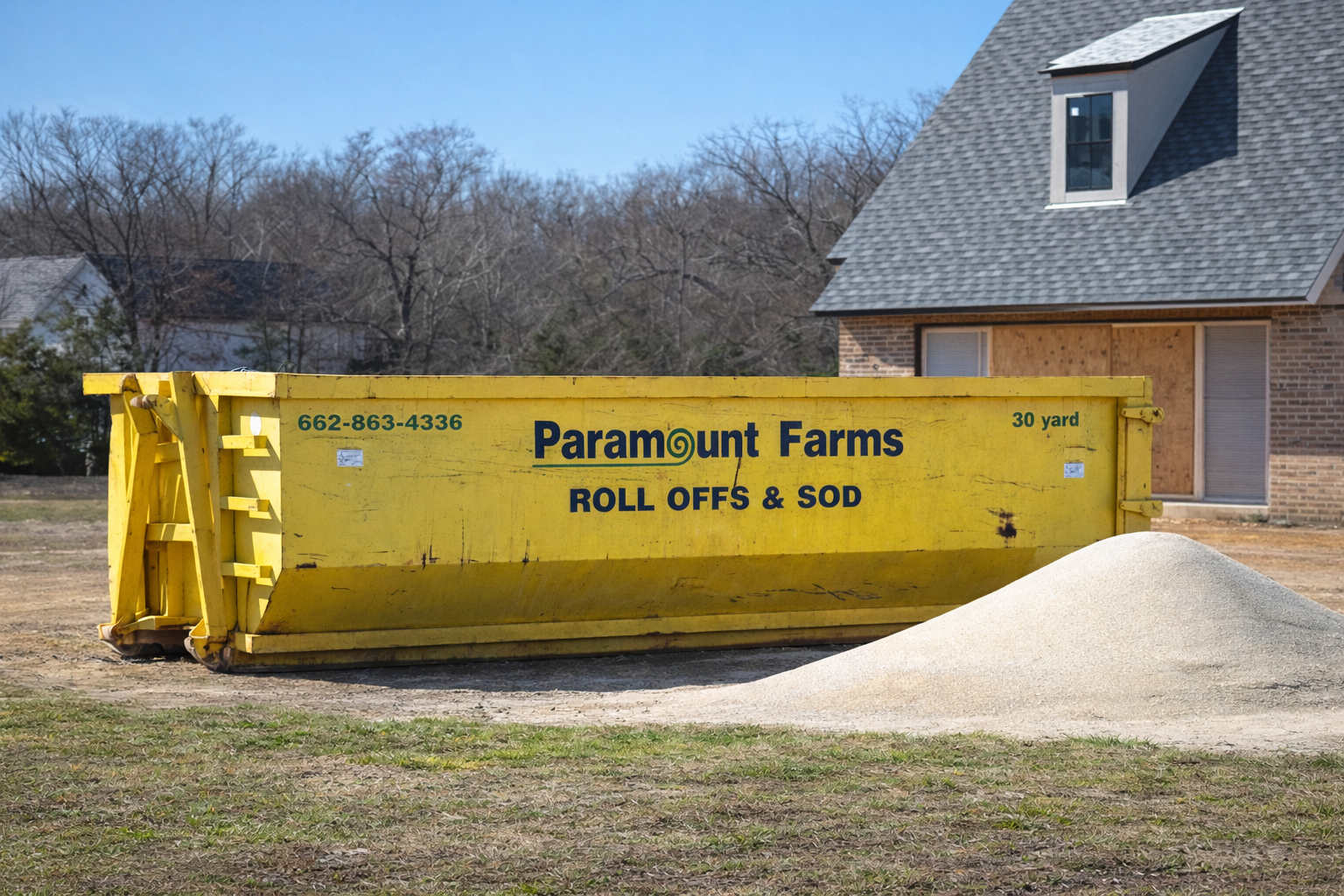 Large yellow dump container filled with sand or gravel on a construction site in front of a partially built house with brick walls and a gray shingled roof.