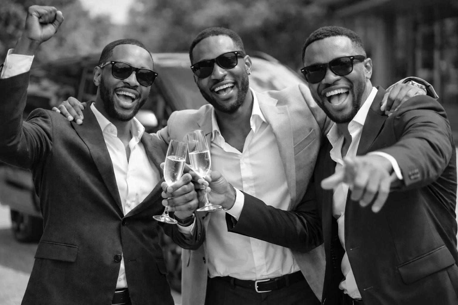 Three men in suits and sunglasses celebrating with champagne, smiling, and raising their glasses in an outdoor setting.