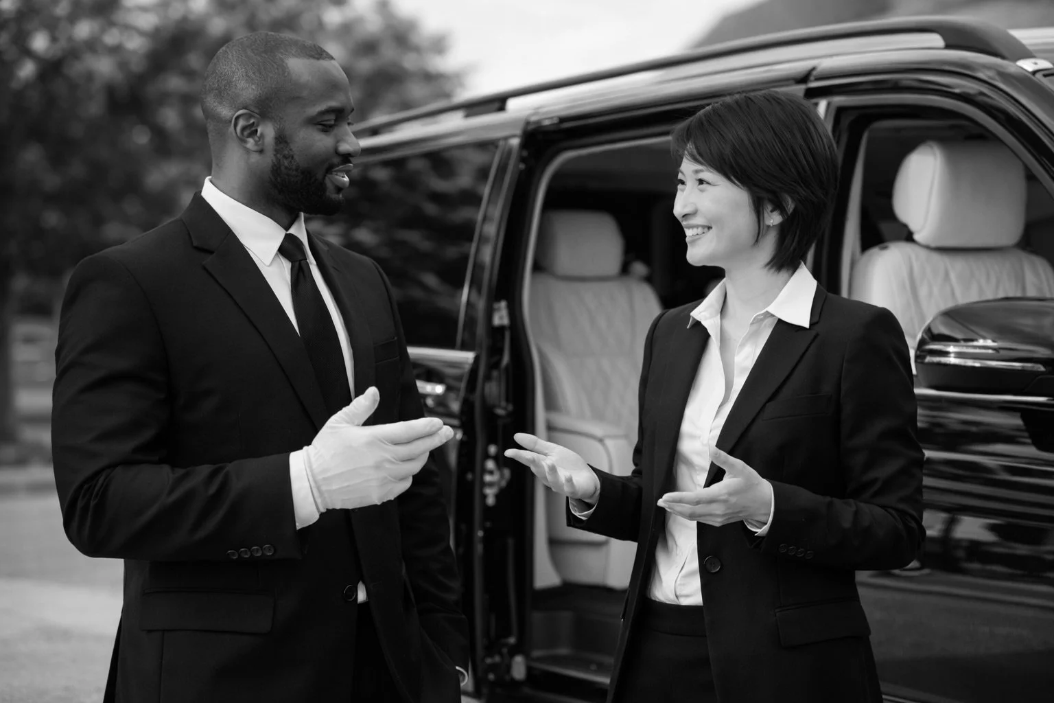 Two professionally dressed people, a Black man and an Asian woman, are standing outdoors near a luxury black vehicle with open doors, engaged in conversation, both smiling.