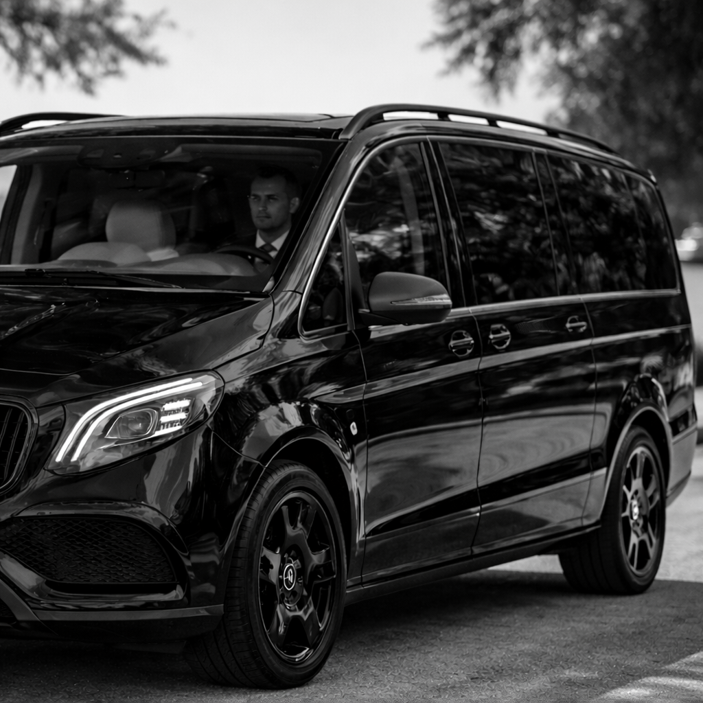 A black luxury minivan parked outdoors with a man in a suit sitting inside, visible through the windshield.