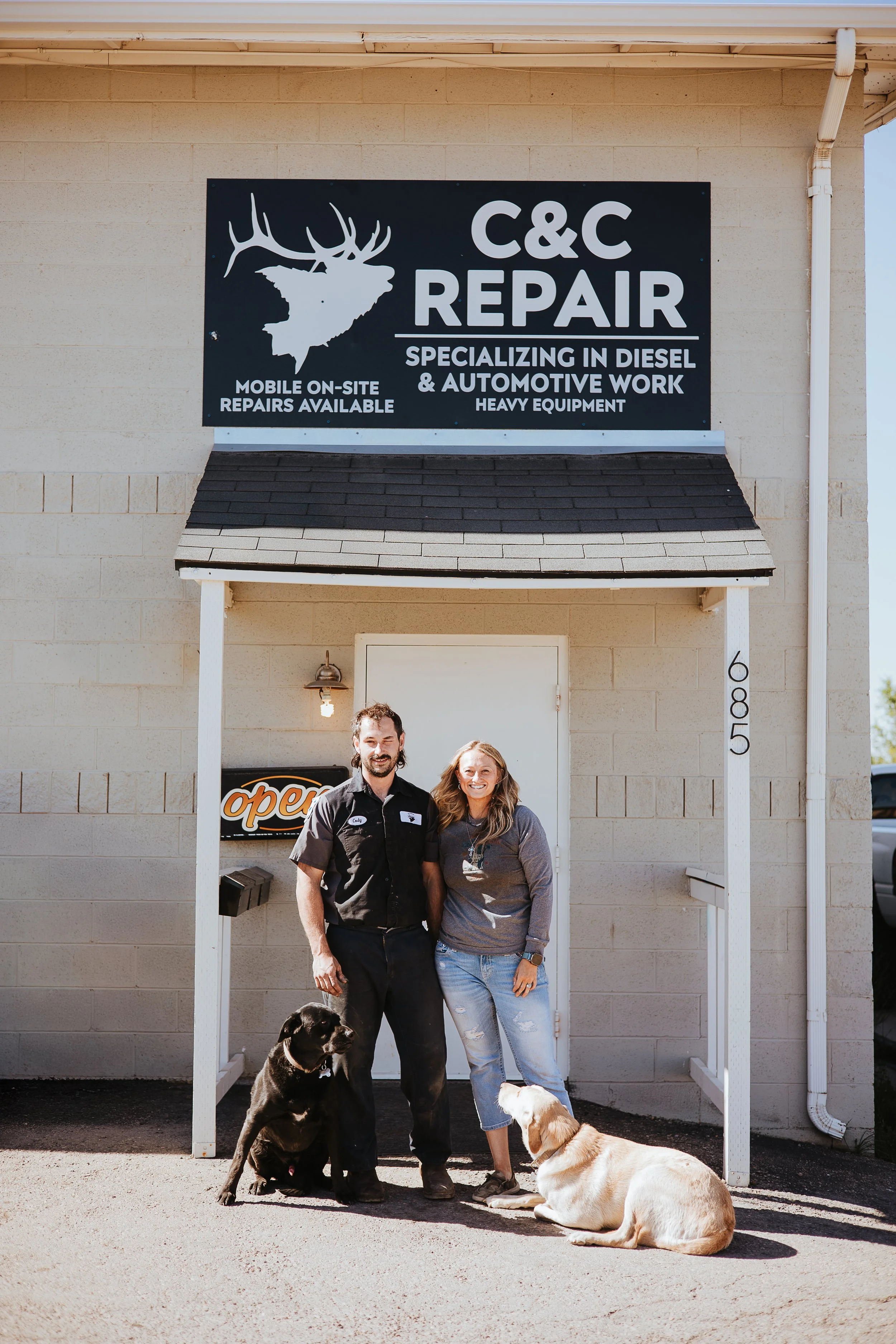 A man and woman standing under a business sign for C&C Repair with animals in front of them, including a black dog sitting, a white and tan dog lying down, and two people.