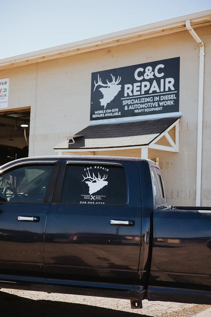 A black pickup truck parked outside a repair shop with a sign that reads 'C&C Repair,' specializing in diesel and automotive work, featuring a logo with a white silhouette of a deer head with antlers.