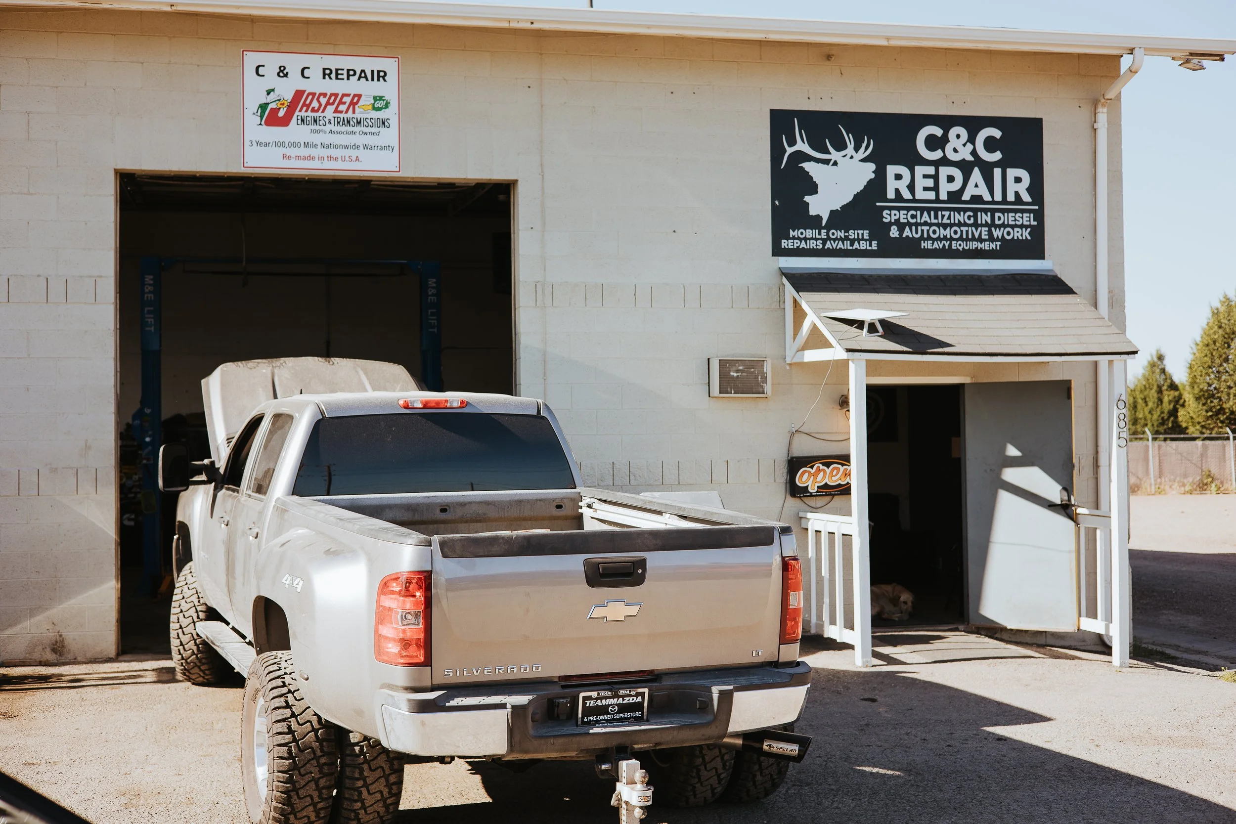 Exterior view of a vehicle repair shop with two signs, one for Jasper Engines and Transmissions and another for C&C Repair, which specializes in diesel and automotive work. A silver Chevrolet Silverado pickup truck is parked in front of the shop, with its hood open and facing away from the camera.