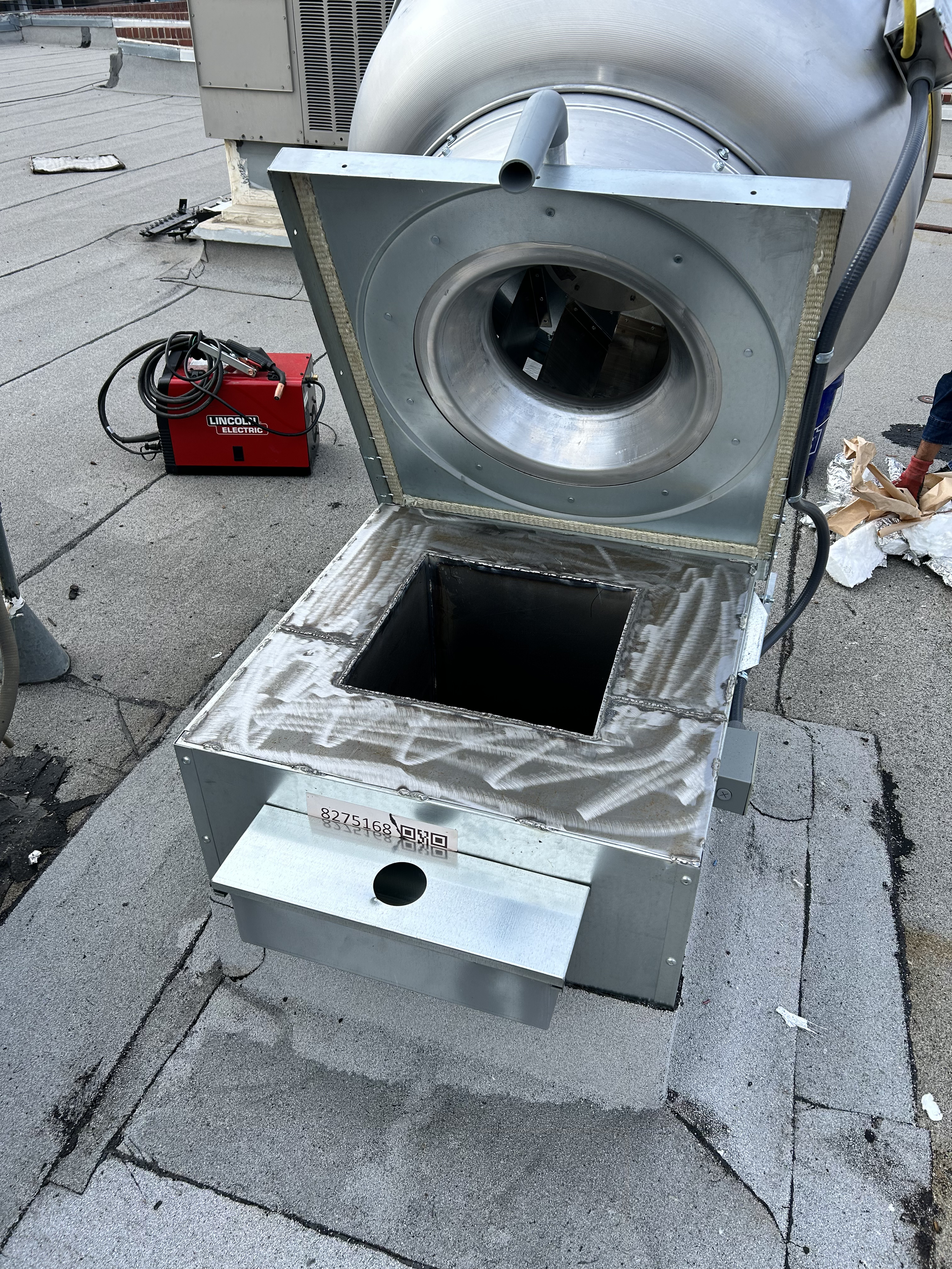 A rooftop HVAC unit with a metal access panel open, exposing the interior ductwork. There is a red welding machine and some tools nearby, indicating ongoing maintenance or installation work.