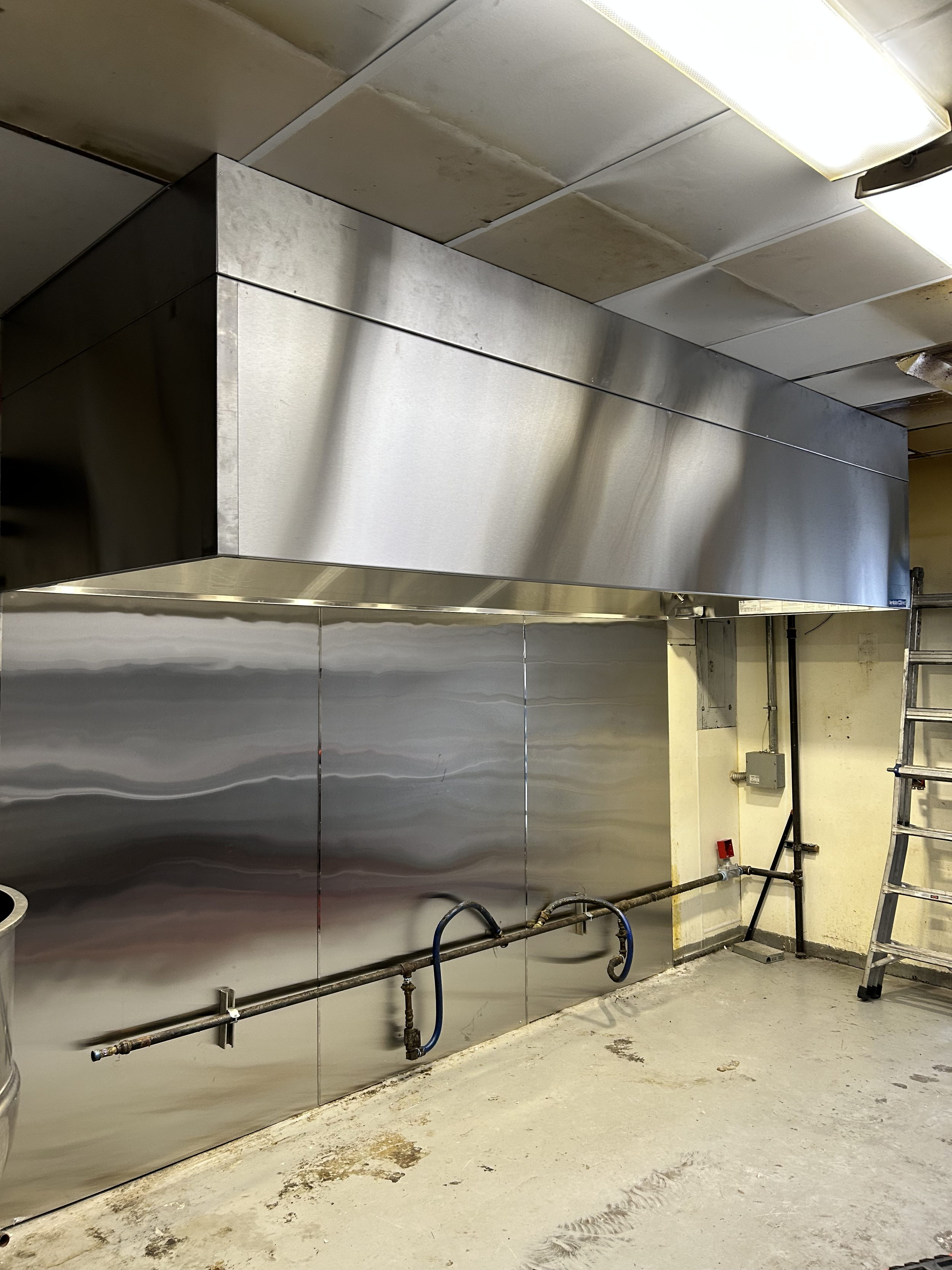 Empty industrial kitchen with stainless steel hood and wall panels, plumbing fixtures, and a ladder against the wall.