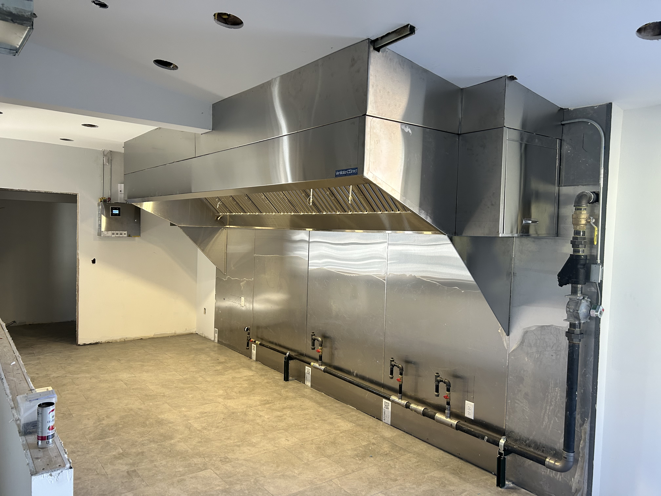 Unfinished commercial kitchen with stainless steel ventilation hood and pipes, beige flooring, and blank walls.