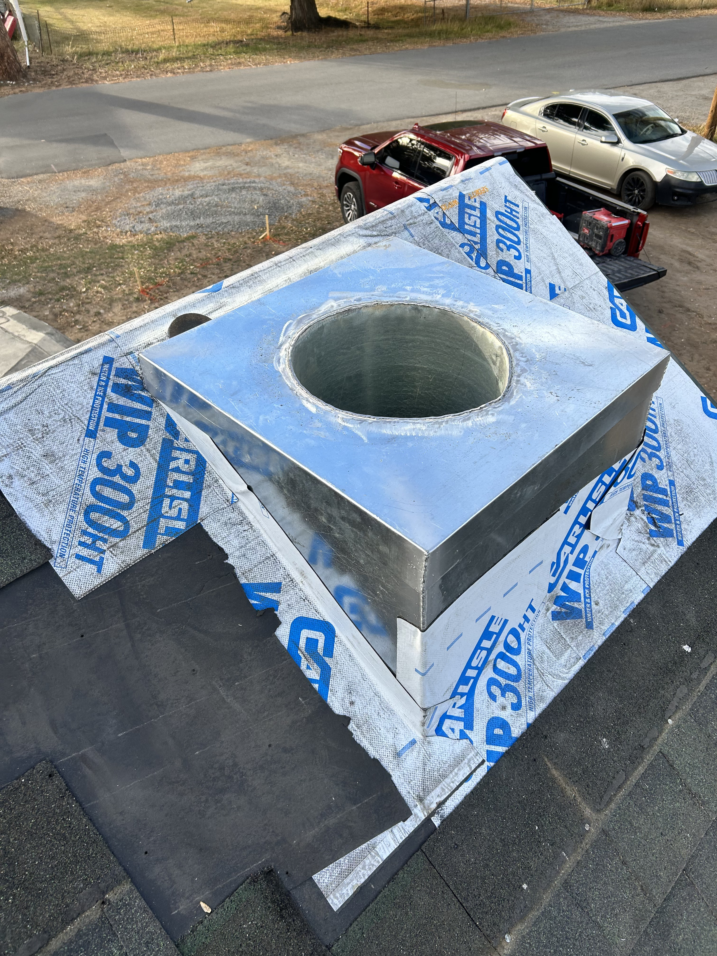 A rooftop vent with a shiny metal cover, surrounded by roofing underlayment marked with blue text, on a building under construction or repair.