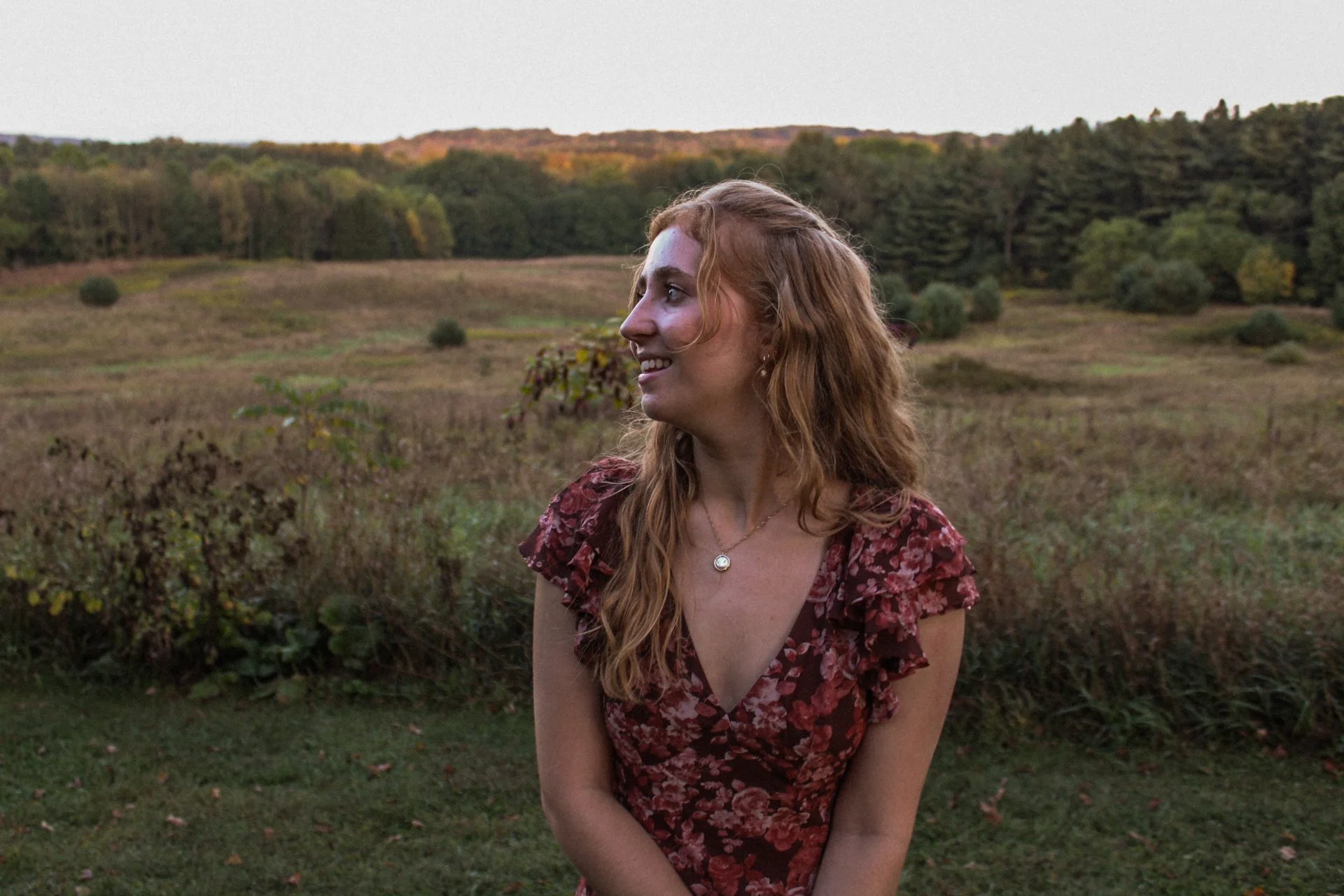 A woman with long red wavy hair standing outdoors in a field with trees in the background, smiling and looking to the side, wearing a red floral dress and a necklace.