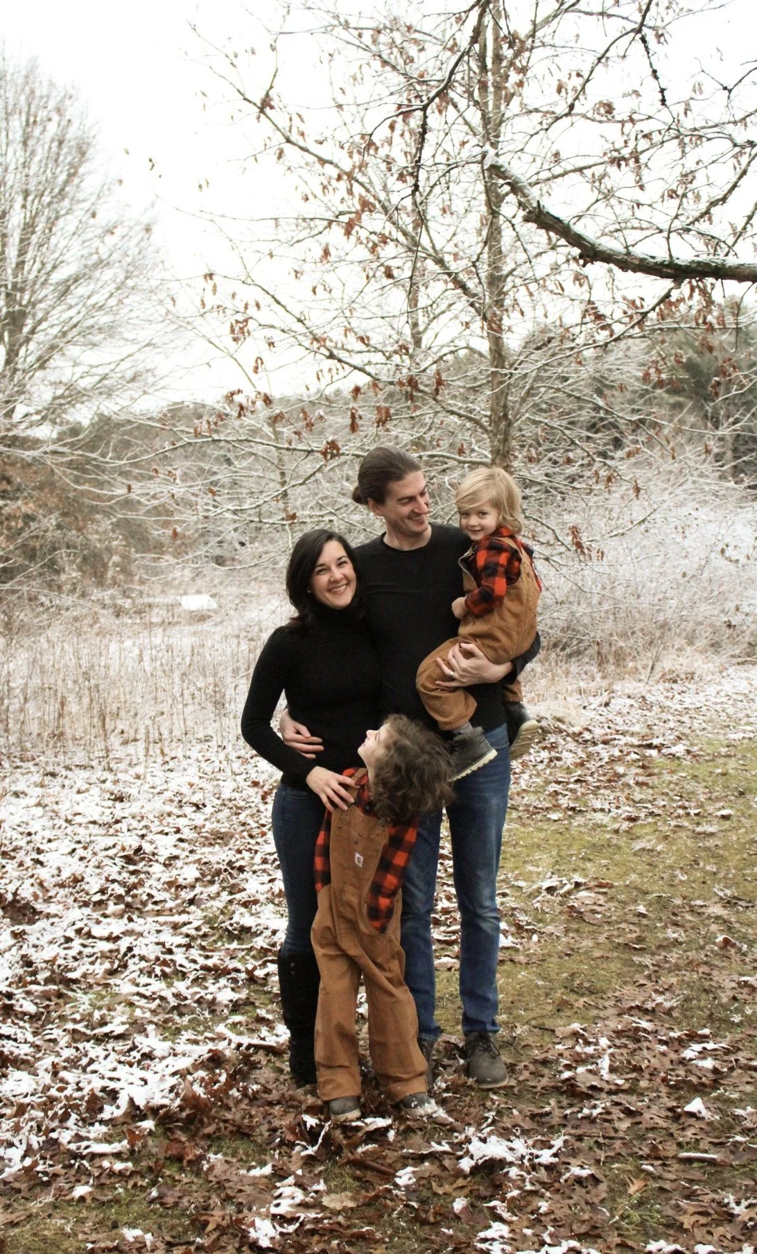 A family of four standing outdoors on a lightly snow-covered ground with leafless trees in the background. The father is holding a little girl, and a woman and a young boy are standing beside him, smiling and looking joyful.