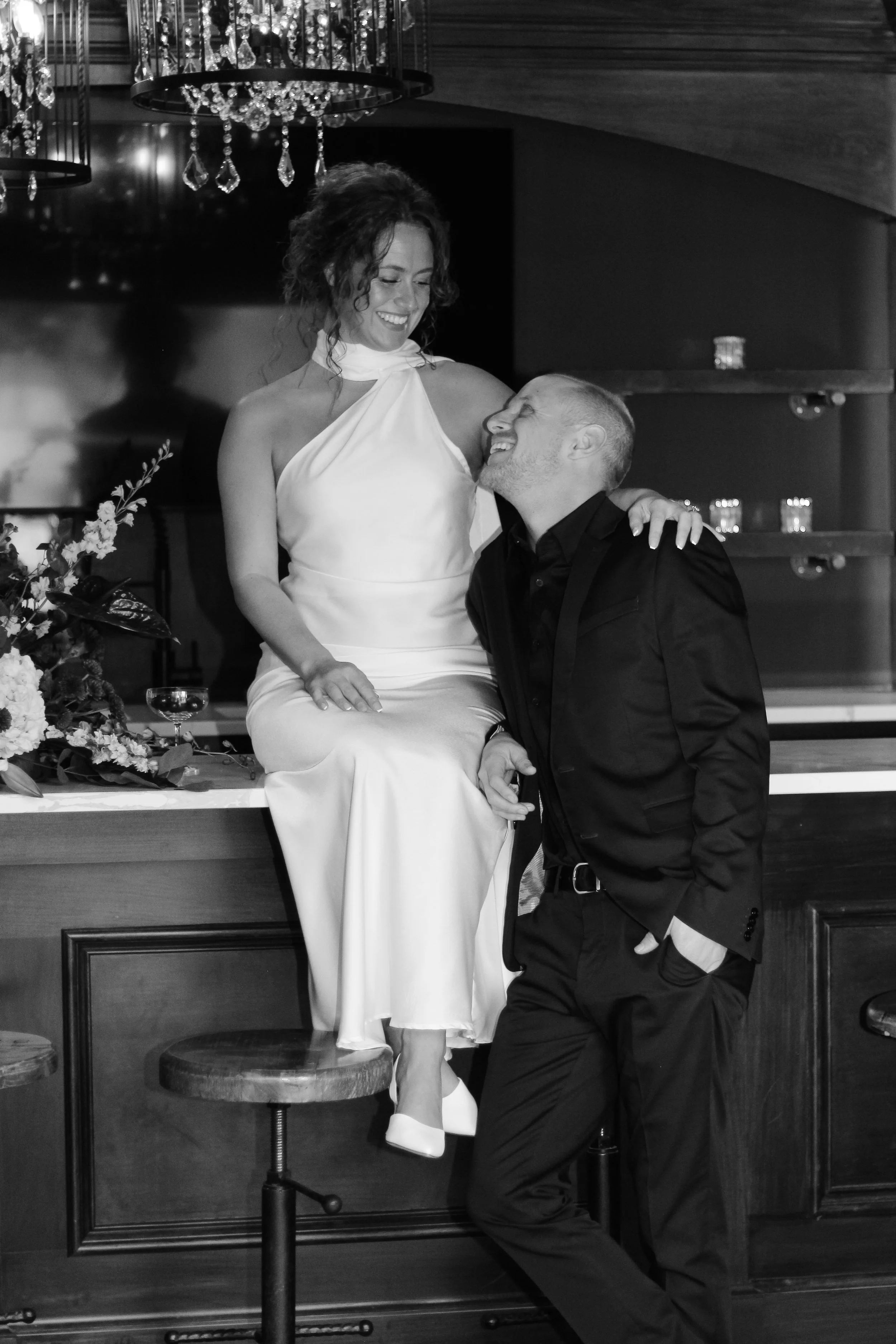 A woman in a white dress sitting on a bar counter, smiling and leaning towards a man in a dark suit, who is standing with one hand in his pocket and the other holding her hand, both sharing a joyful moment in a dimly lit setting.