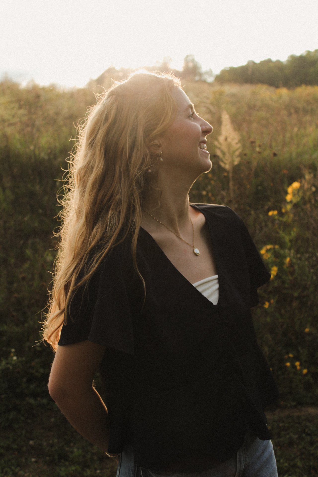 A woman with long wavy blonde hair smiling and looking to the right, outdoors during sunset, wearing a black top with a white inner layer and a gold necklace, with trees and hills in the background.
