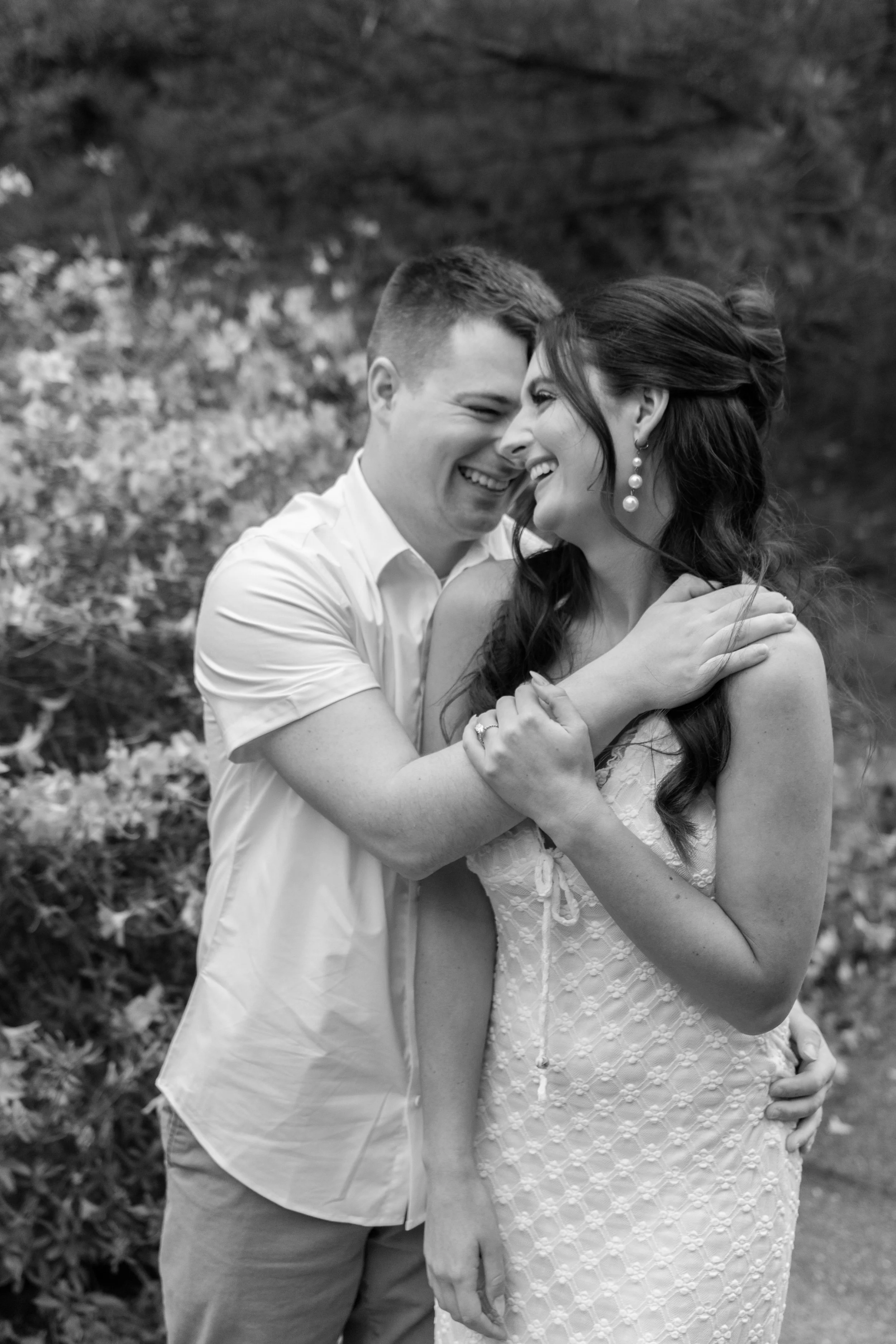 A joyful couple embracing outdoors, showing a close-up of their faces and upper bodies. The man is wearing a light-colored shirt and the woman is wearing a sleeveless dress with a textured pattern and large pearl earrings. They are smiling and touchi