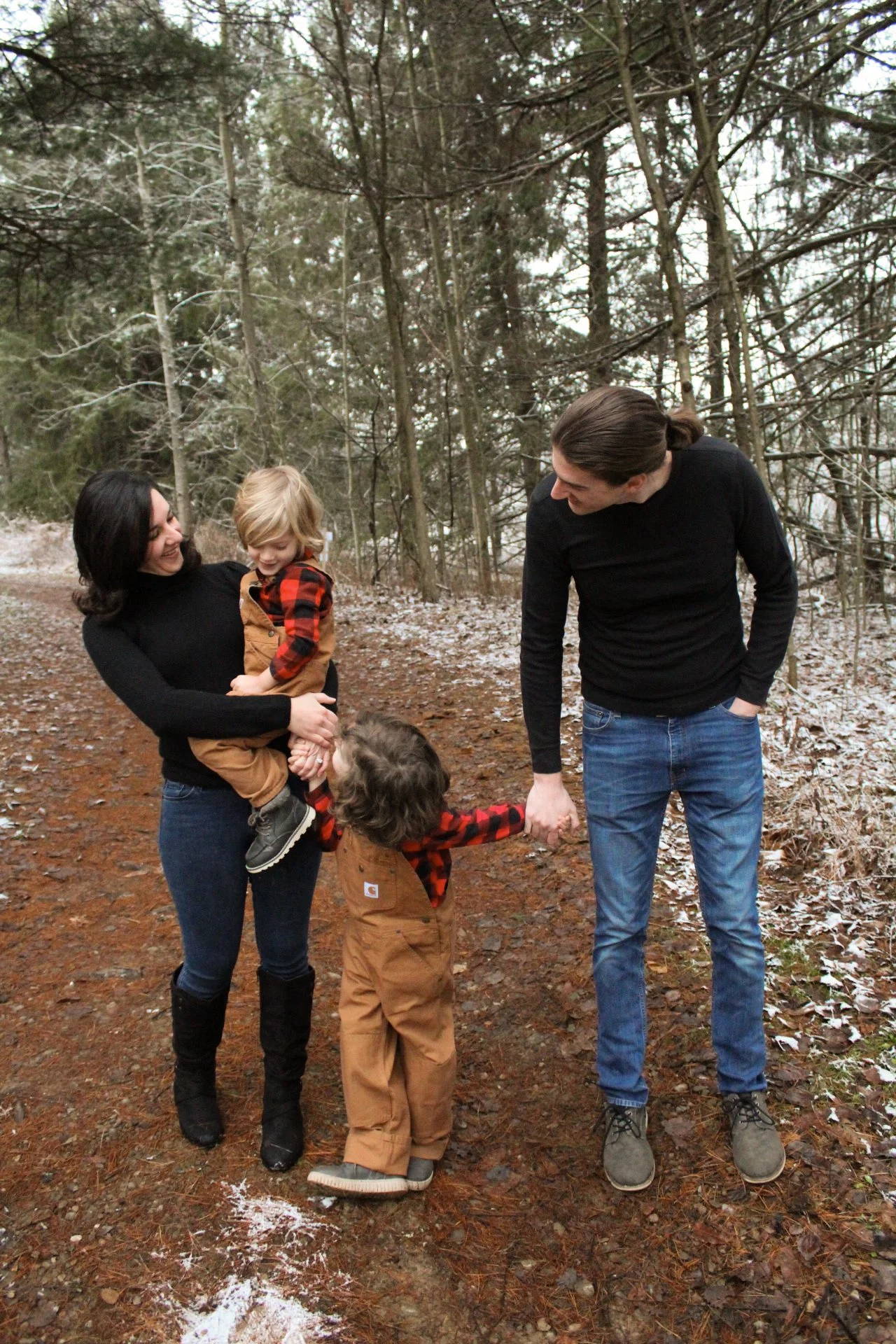 Family of four enjoying a walk in the woods during winter; mother holding young son, father holding daughter's hand, all smiling.