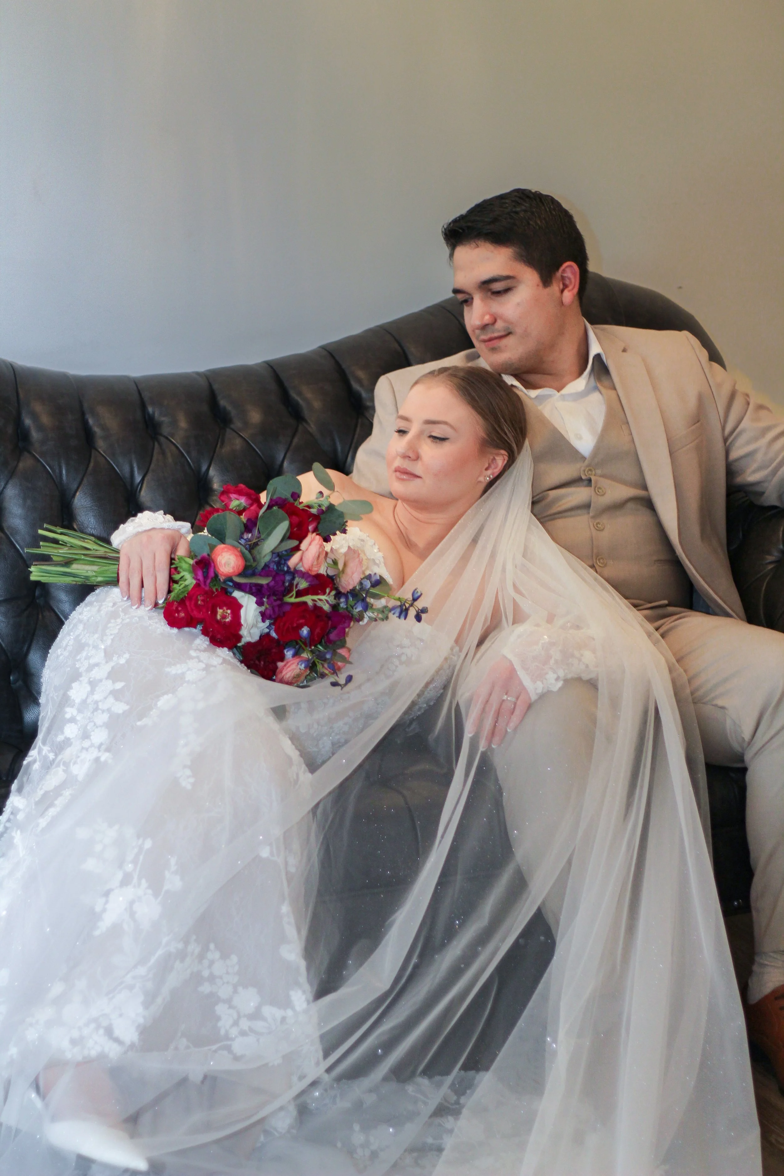 A bride and groom sitting on a black leather couch, with the bride resting her head on the groom's shoulder. The bride is holding a large bouquet of red, pink, purple, and green flowers, wearing a white lace wedding dress and veil. The groom is dress