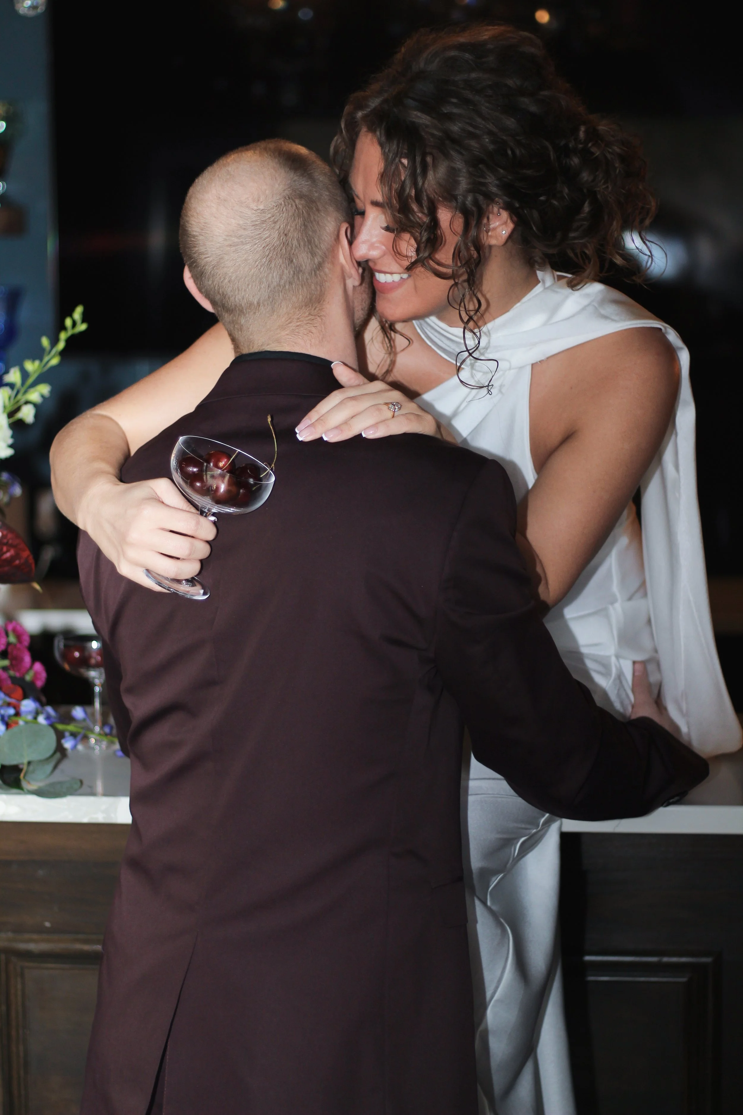 A man and woman embrace closely at a celebration, with the woman holding a glass of cherry. The woman is smiling, and they appear happy.