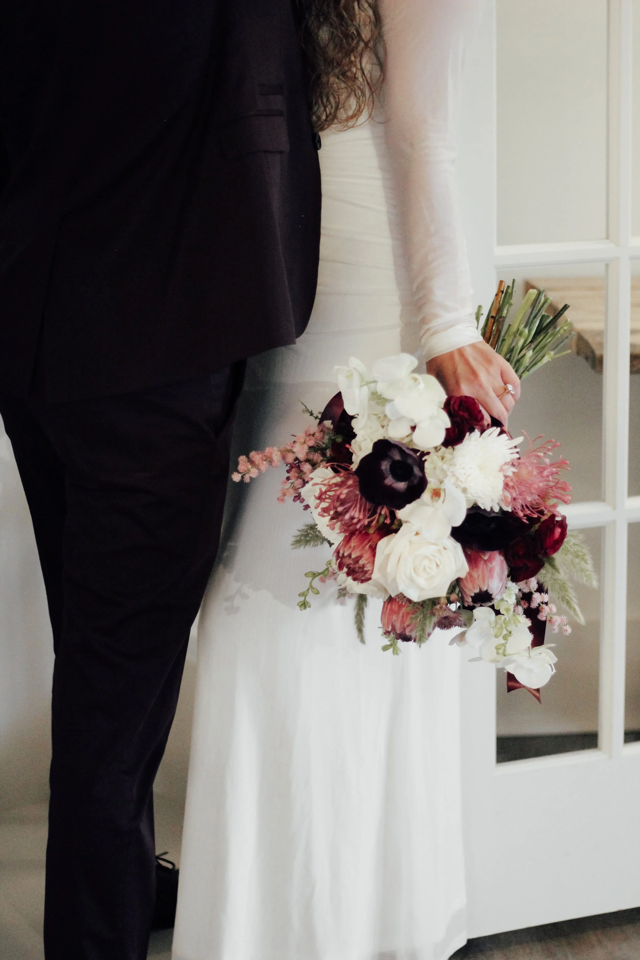 A bride holding a bouquet of flowers, standing next to a person in a tuxedo. The bouquet includes white, red, and dark purple blooms, with some greenery.