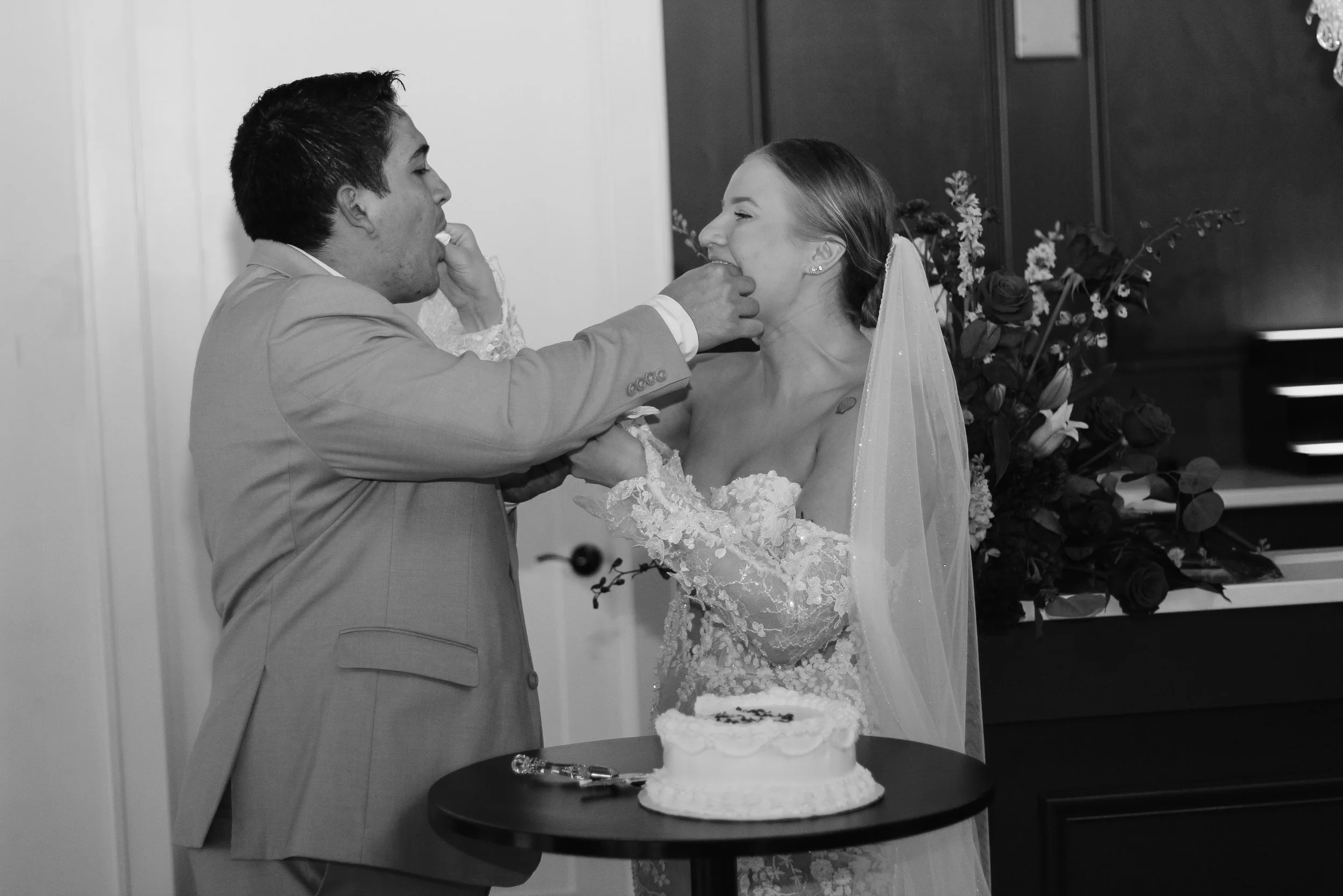 A black and white photo of a wedding reception with a couple feeding each other cake. The bride is smiling with her eyes closed, wearing a lace wedding dress and veil. The groom is dressed in a suit, with a hand on her face. A wedding cake is on a sm