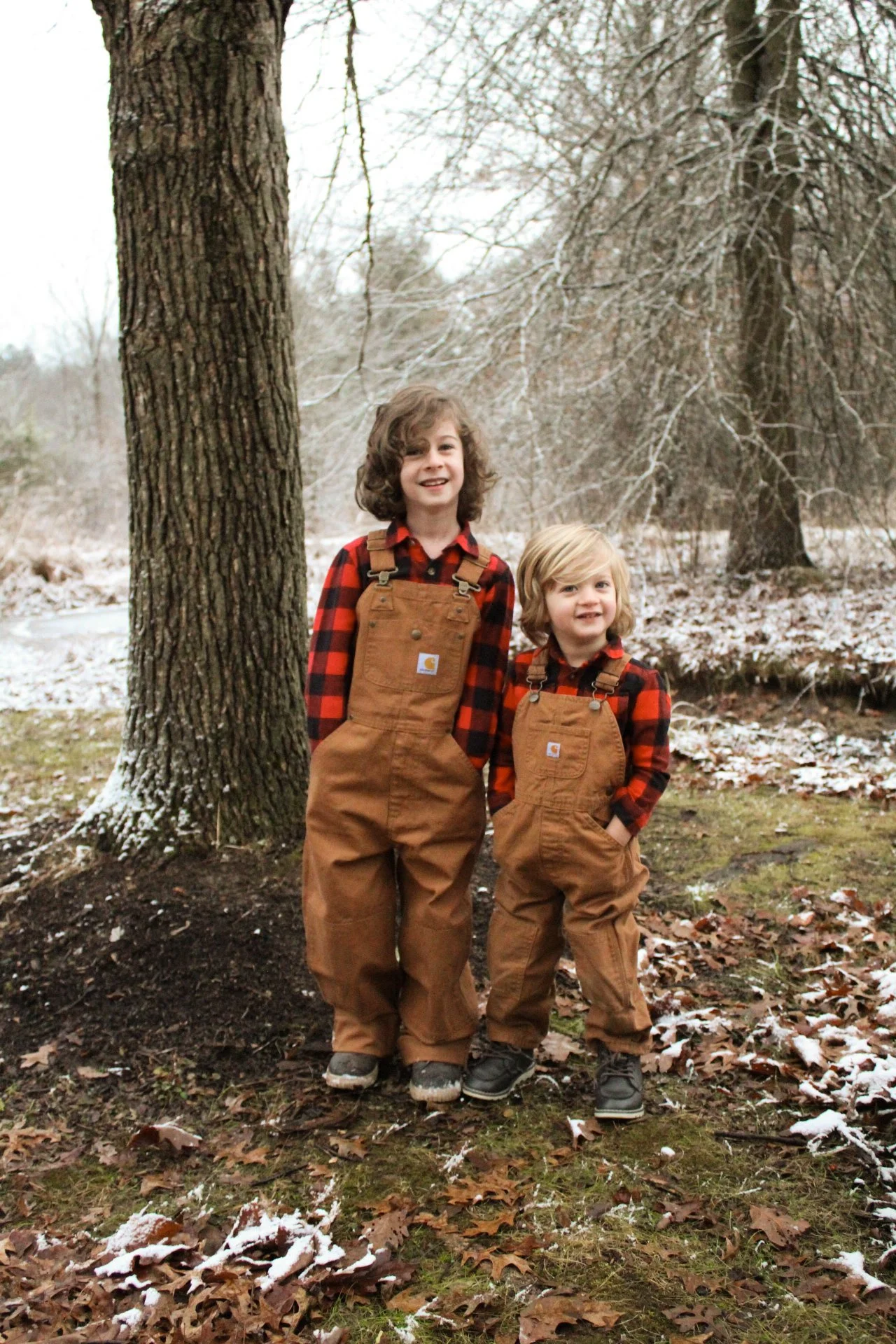 Two young boys, wearing brown overalls and red plaid shirts, stand outdoors in a wooded area with fallen leaves and snow. They are smiling and posed near a large tree.