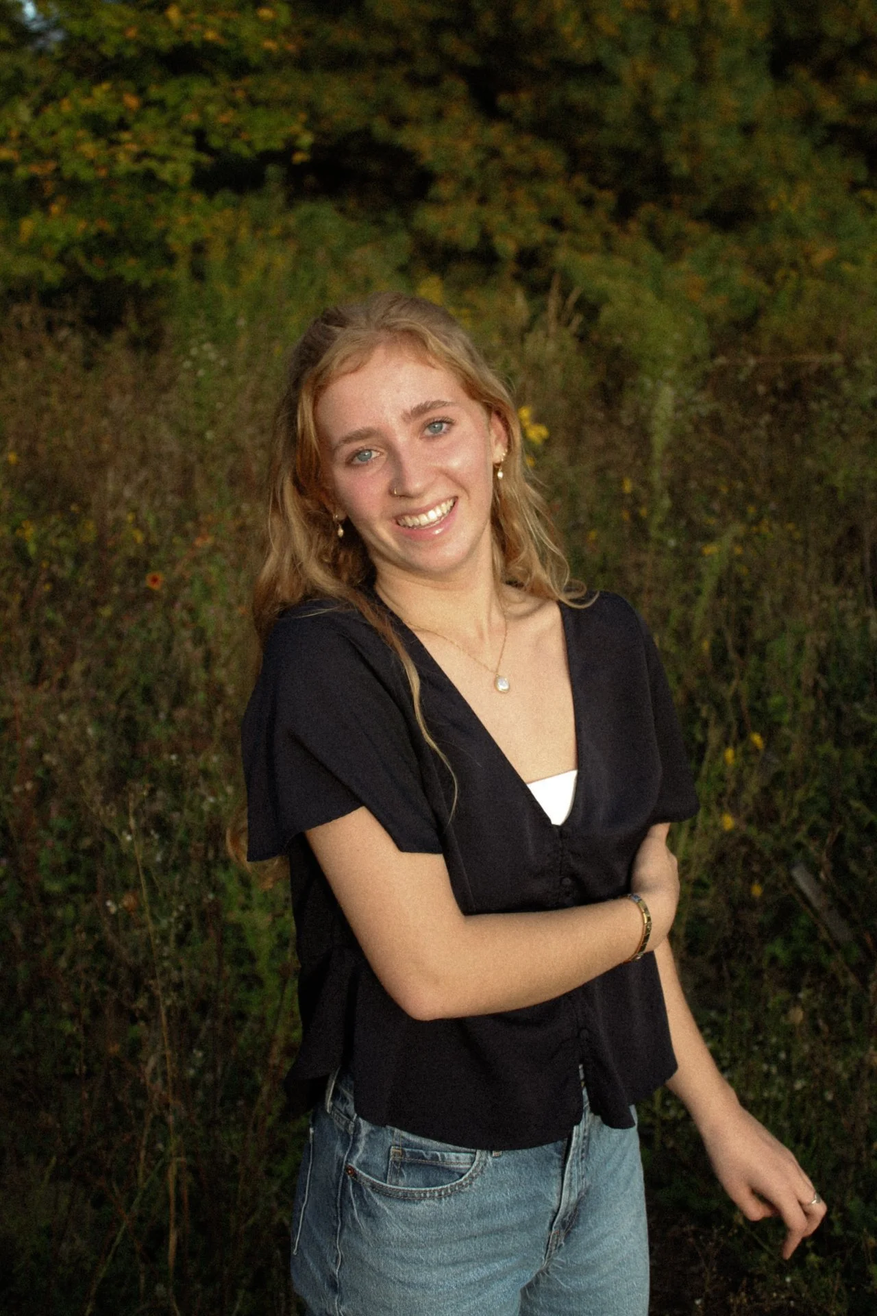 A young woman with curly blonde hair and blue eyes smiling outdoors in a wooded area, wearing a black shirt, jeans, jewelry, and a white top underneath.