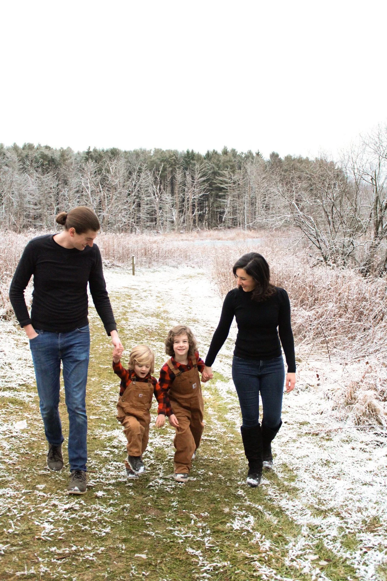 A family of four walking on a snowy trail in winter; the father holding the younger child's hand, the mother holding the older child's hand, all smiling and enjoying the outdoors.
