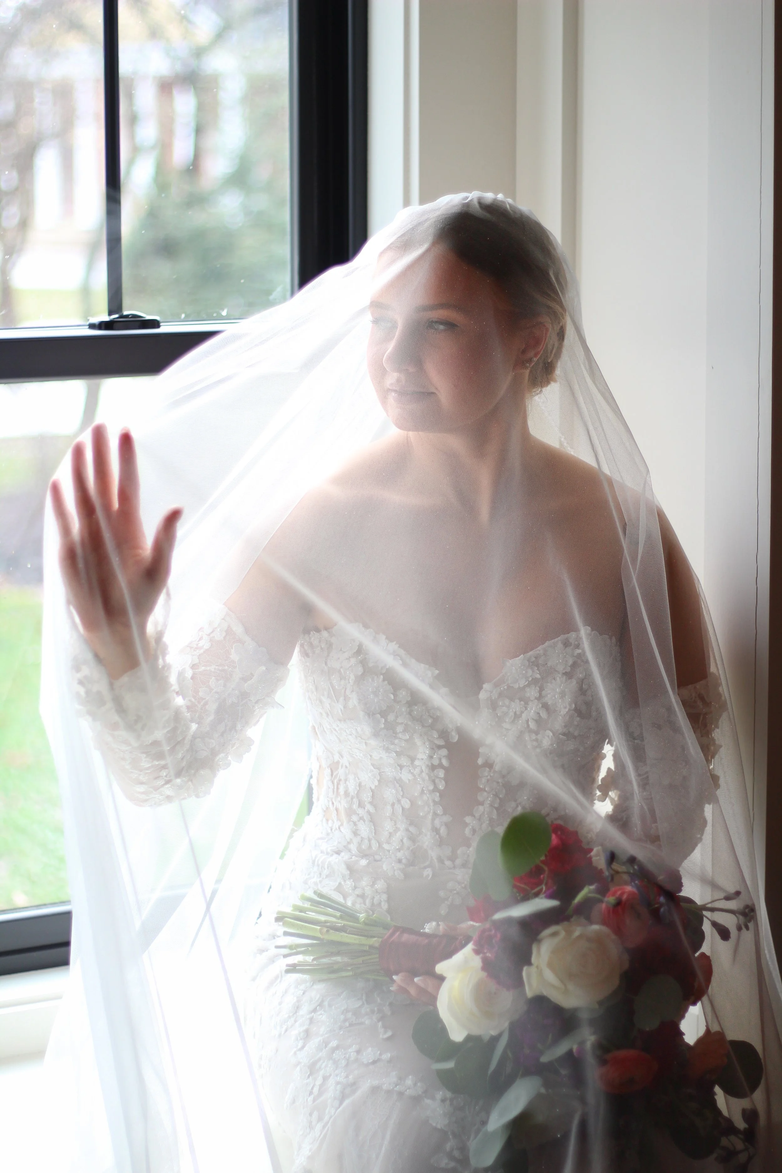 Bride in a white wedding gown and veil holding a bouquet, standing near a window and touching the glass.