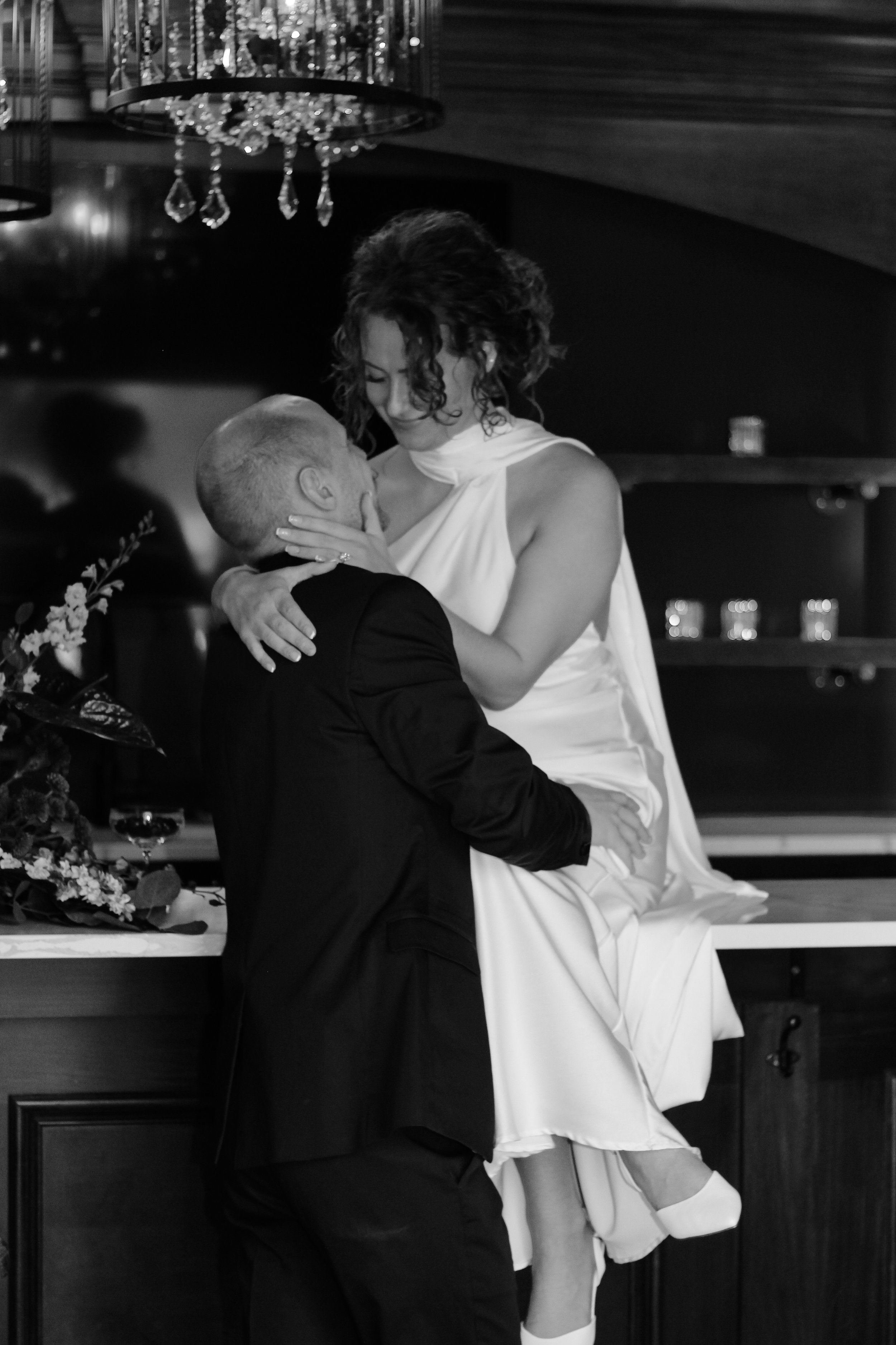 A woman in a white dress sitting on a kitchen counter, being held and kissed by a man in a suit, with a chandelier overhead and a floral arrangement nearby.