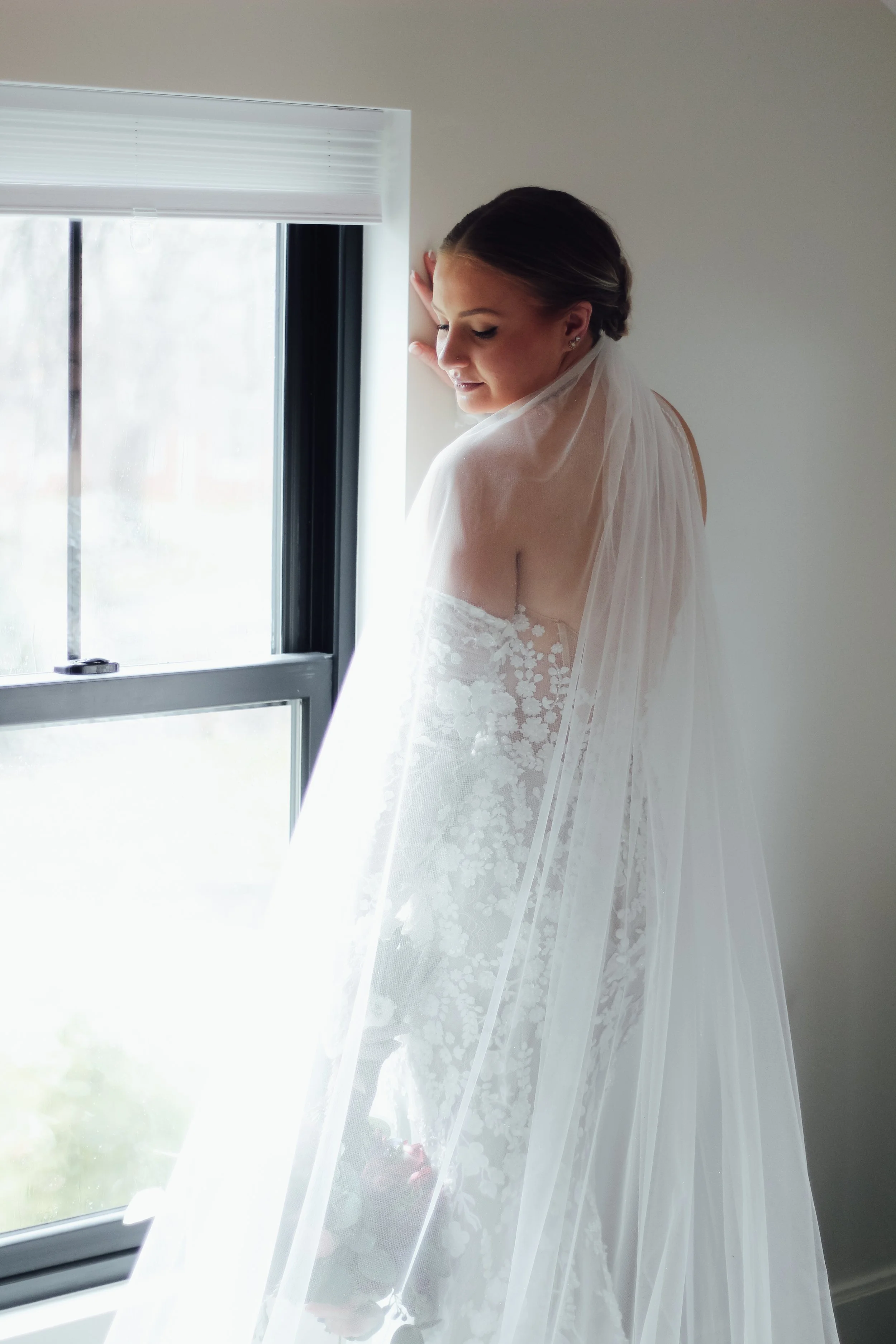 A bride in a white wedding dress with floral lace details looks down and leans against a window, with daylight coming through the window and illuminating her face.
