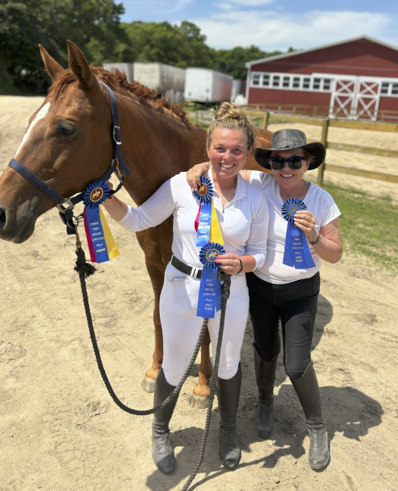 Professional horse trainer Emma Davern with a show client. Two women and a horse at a dirt arena, holding blue and yellow award ribbons, with a red barn and green trees in the background.