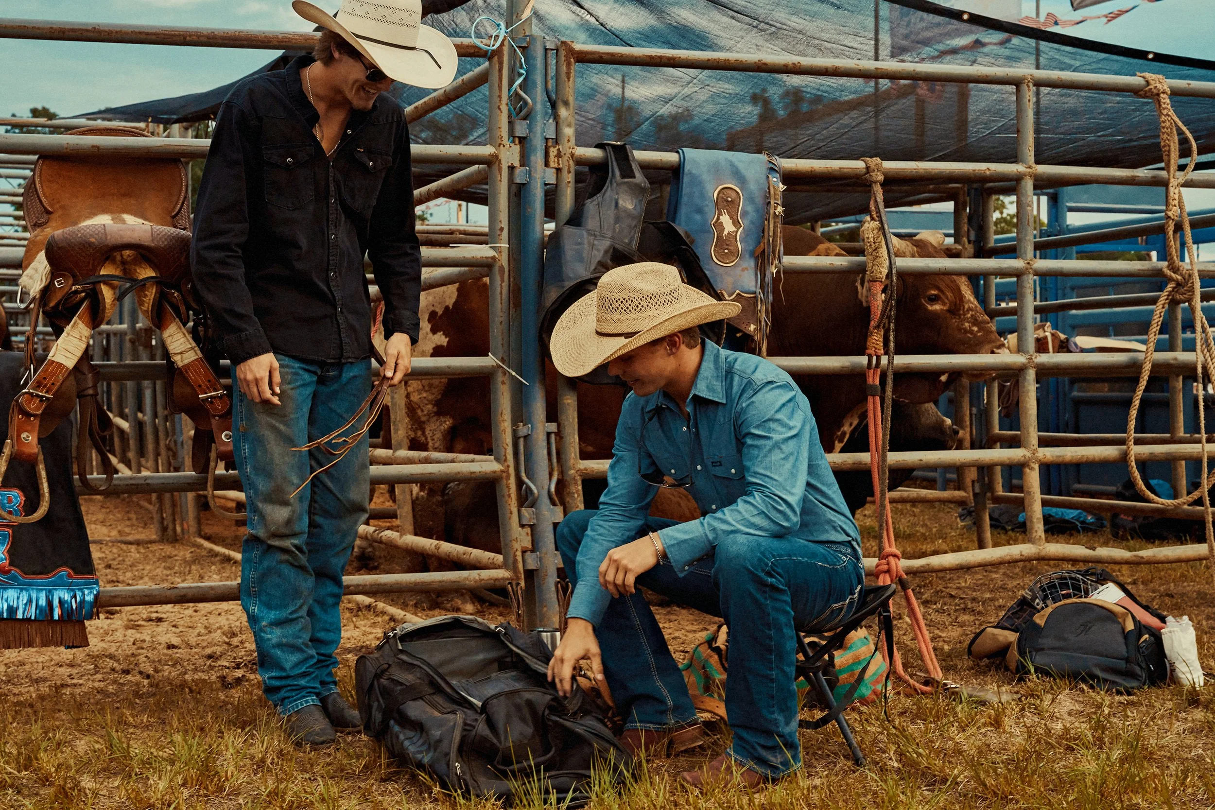 Two men at a cattle ranch, one standing and one sitting, packing bags near cattle pens with cows in the background.