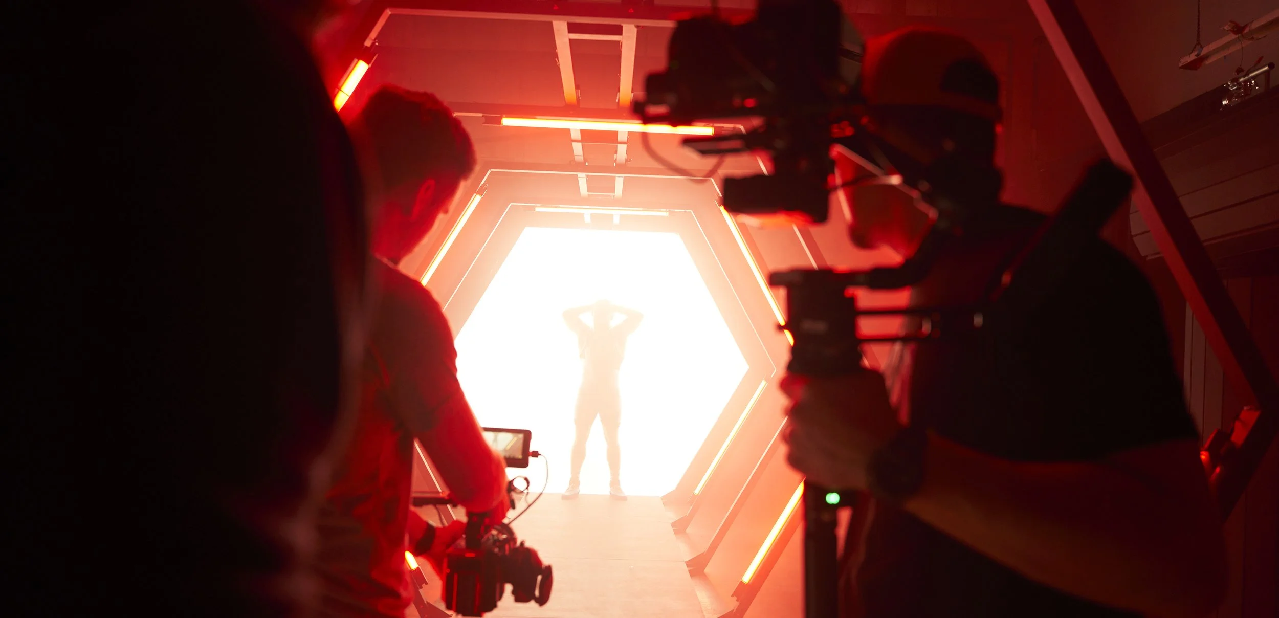 People filming a person standing with hands behind head inside a brightly lit hexagonal tunnel with red lights.