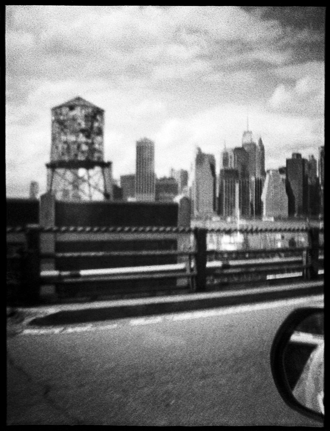 Black and white photo of a city skyline with tall buildings, a water tower in the foreground, and a road with a side mirror visible on the right.