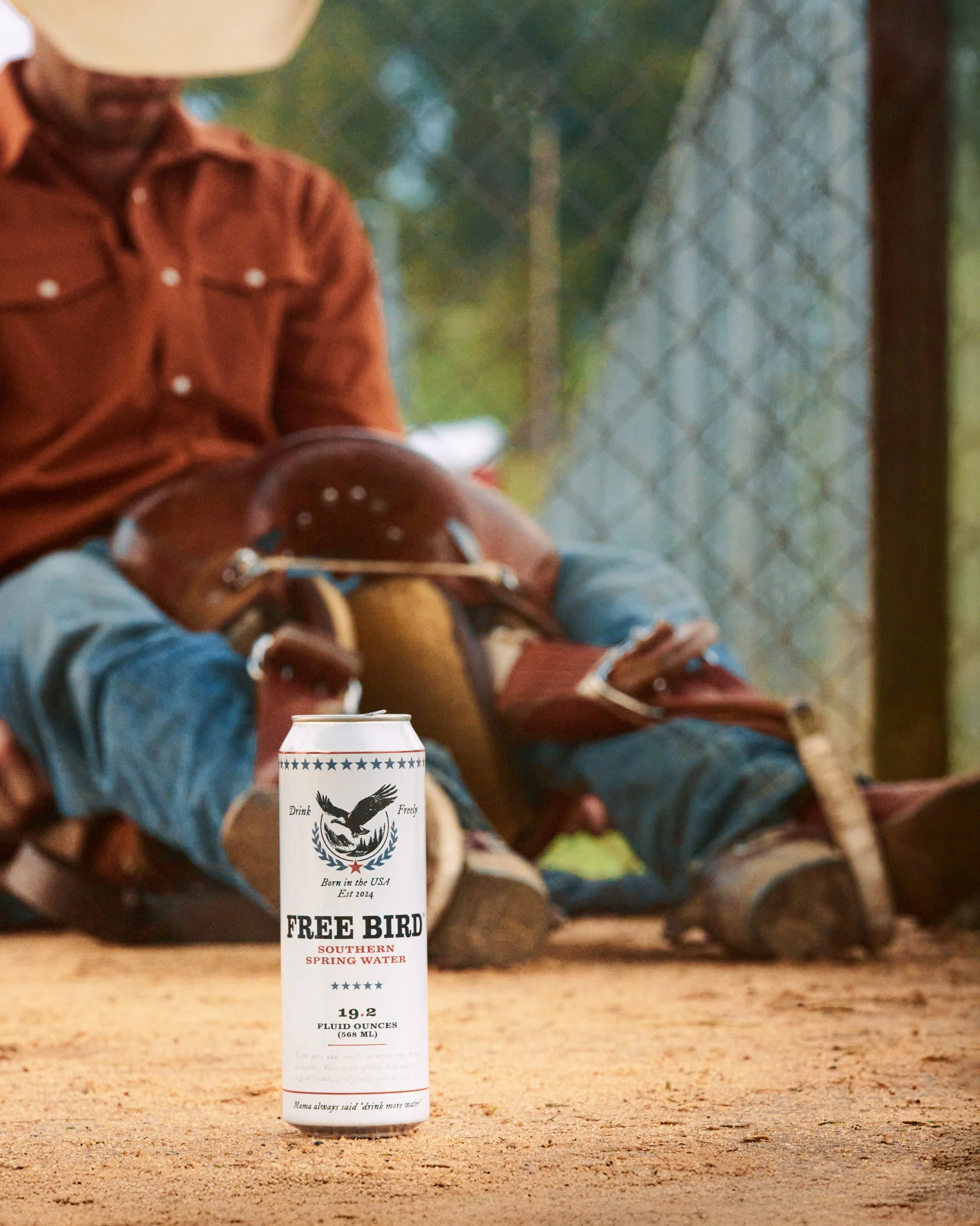A can of Free Bird Southern Spring Water placed on the ground in front of a horse rider sitting on the ground with a helmet, cowboy hat, and boots, with a chain-link fence and trees in the background.