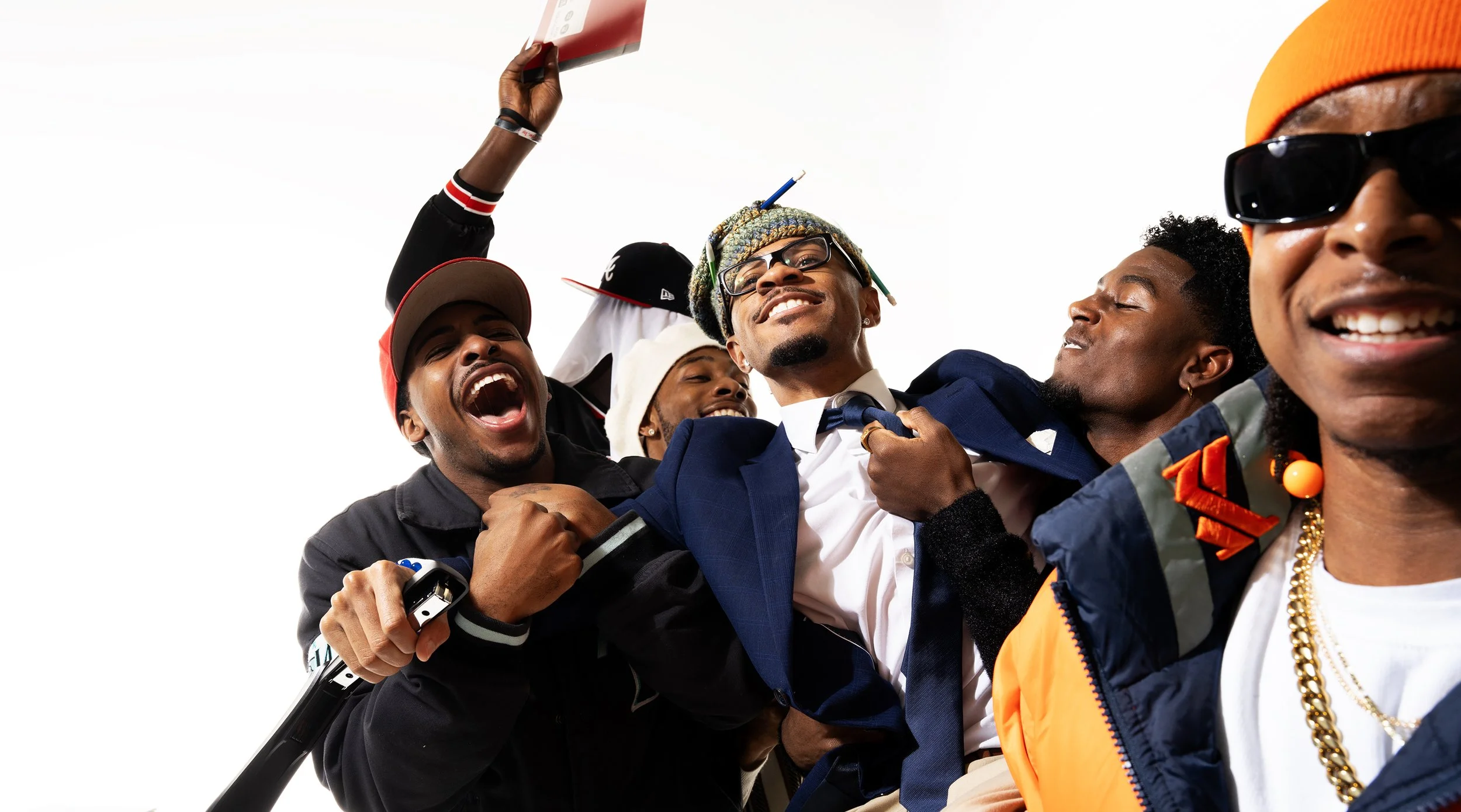 Group of young African American men smiling and celebrating together, with one man in the center wearing glasses and a colorful beanie, dressed in a suit and tie, surrounded by friends.