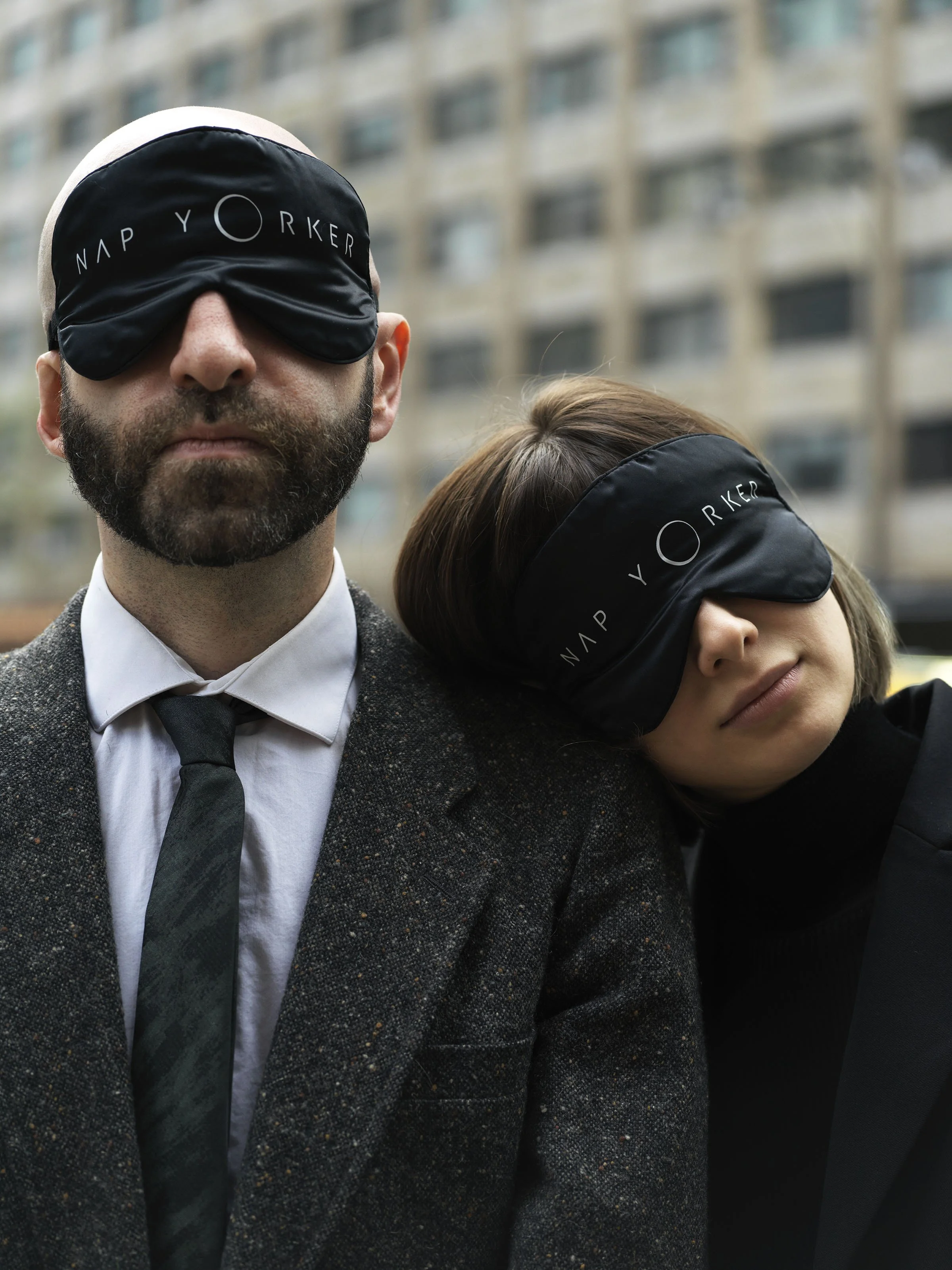 A man and woman standing outdoors in front of a building, both wearing black sleep masks with the words 'NAP Y O R K E R' on them. The man is wearing a suit and tie, and the woman is resting her head on his shoulder.