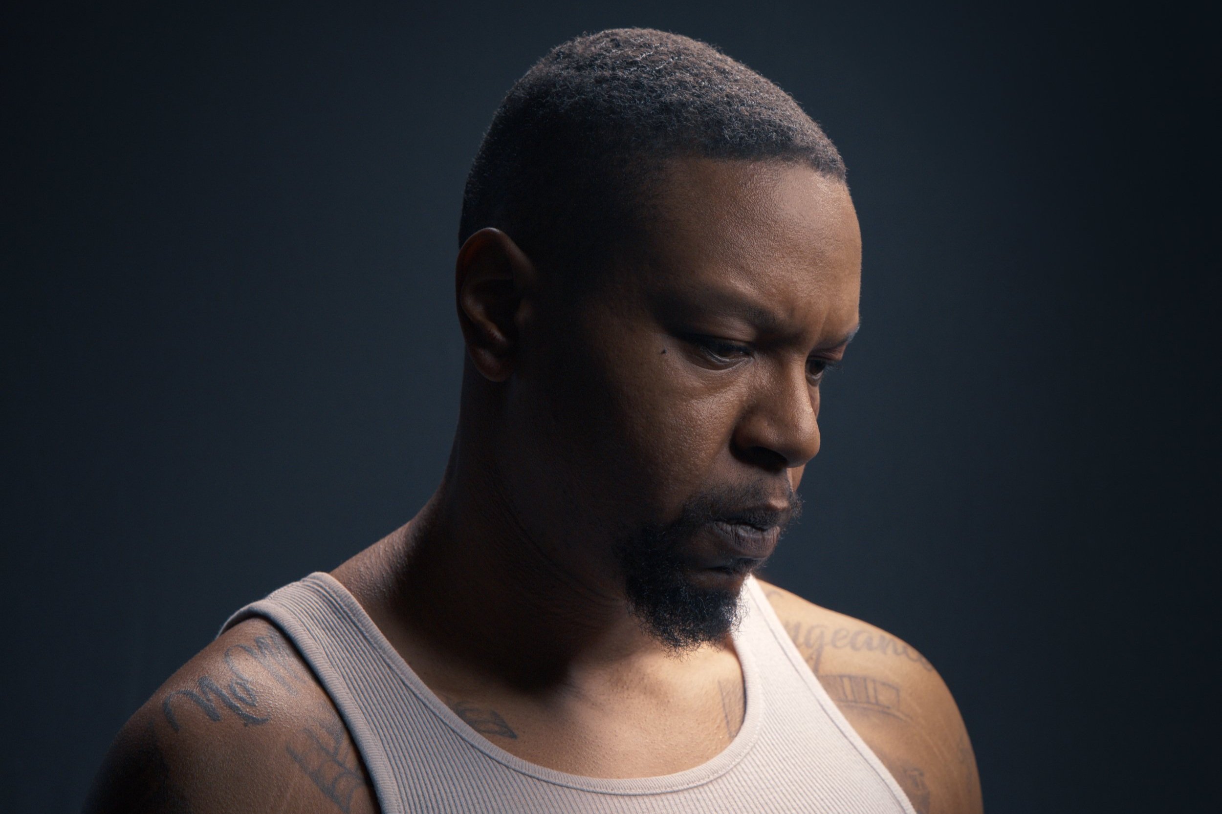 A man with short hair and tattoos on his shoulder looks downwards with a serious expression, wearing a light-colored tank top against a dark background.