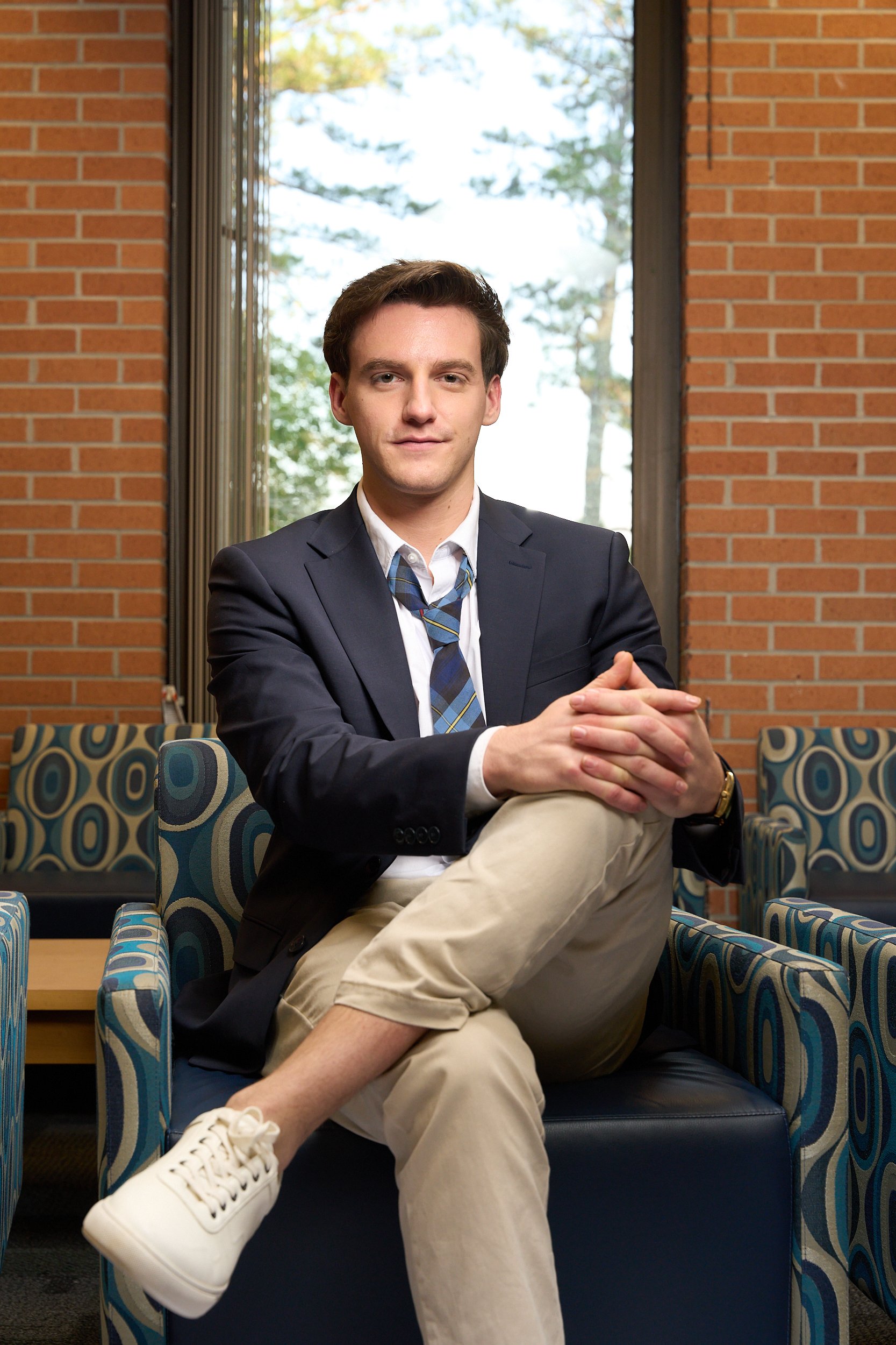 A young man in a navy blazer, white shirt, and striped tie sitting on a modern sofa in a room with brick walls and large window behind him, with trees visible outside.