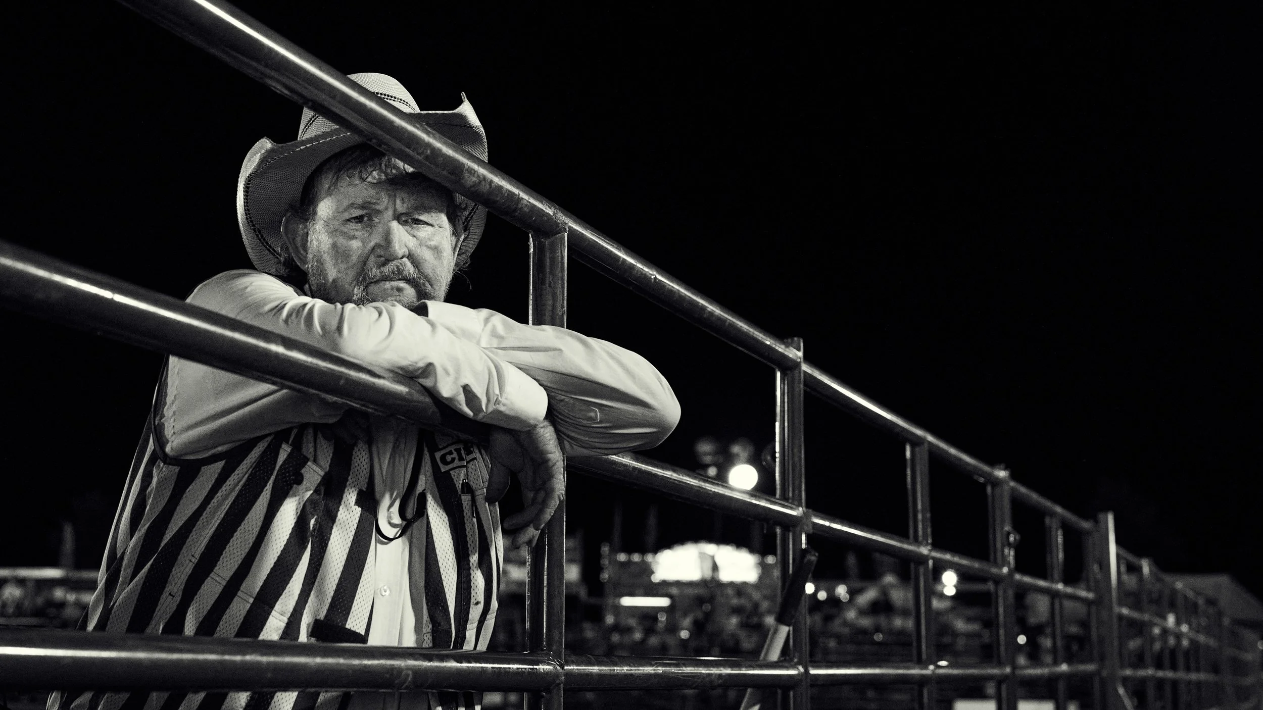 A man wearing a cowboy hat and striped shirt leans on a metal railing at night, looking directly at the camera with a serious expression.