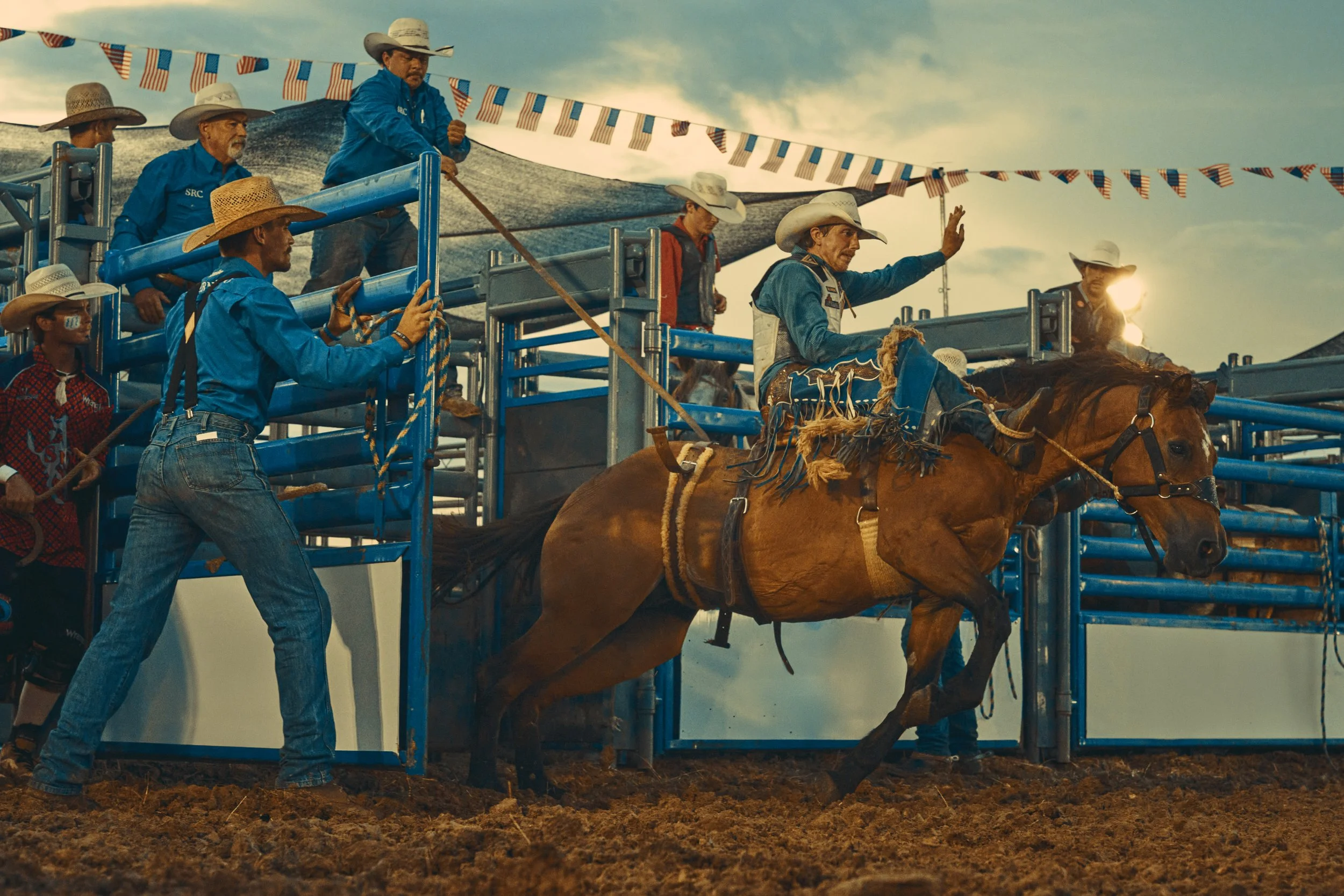 A rodeo event with a cowboy riding a bucking horse, surrounded by rodeo personnel and spectators, with a cloudy sky in the background.