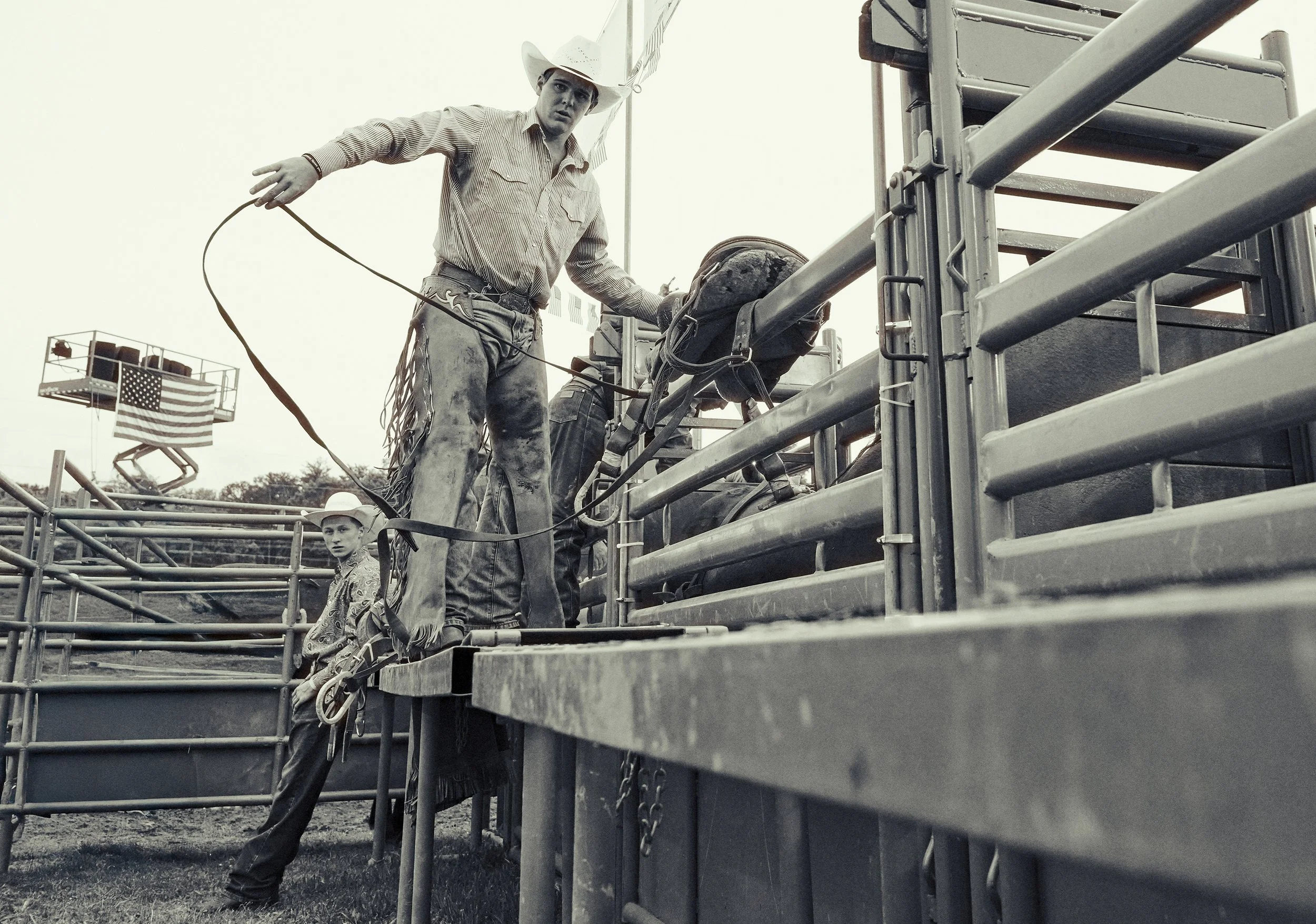 Two cowboys in cowboy hats and jeans working getting ready to compete in bronc riding at a rodeo, with an American flag in the background.
